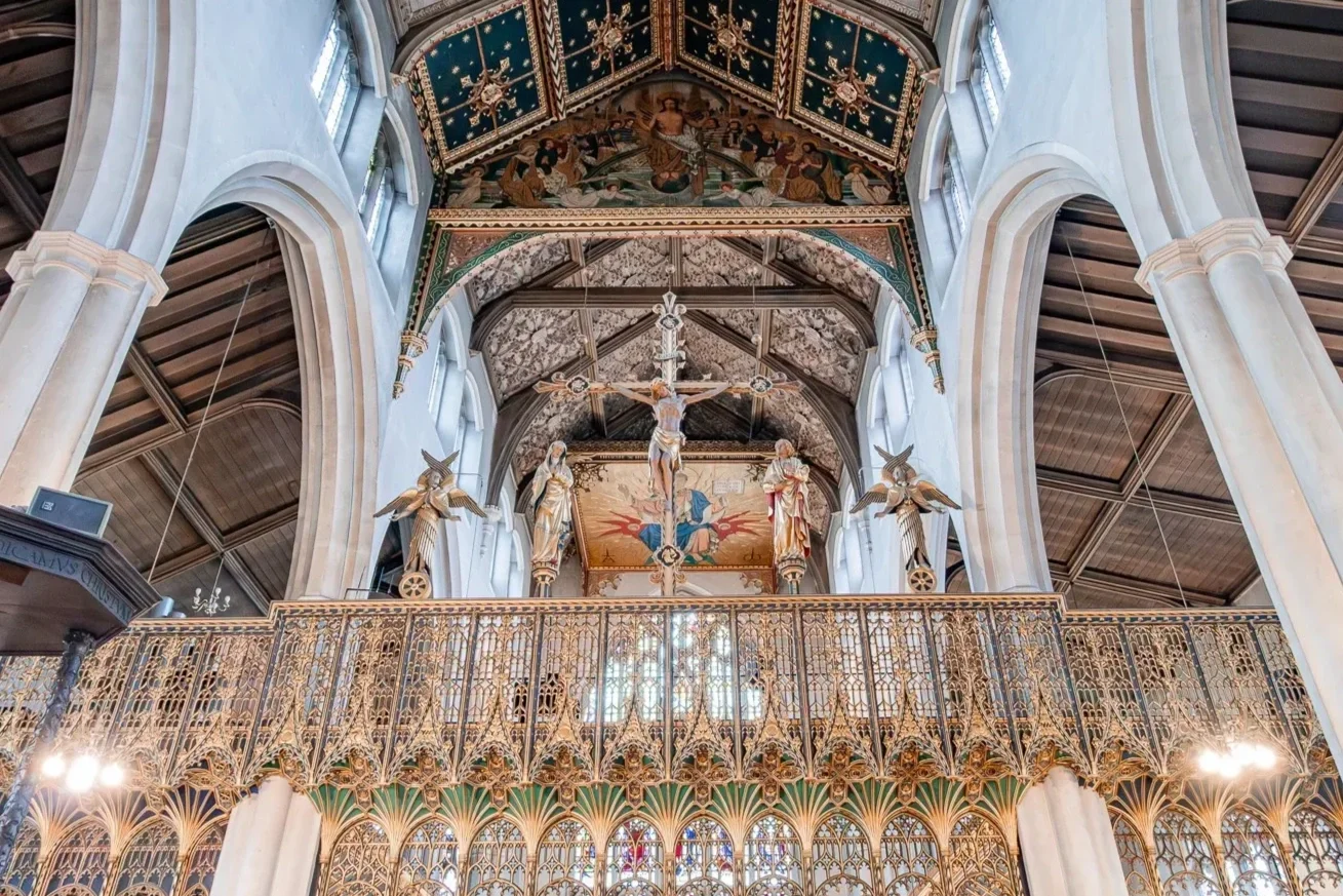 Interior of a church with a high vaulted ceiling, large stained glass windows, and ornate gold railing. There are religious sculptures and paintings, including a crucifix with Jesus on the cross and figures of saints, along with decorative ceiling artwork.