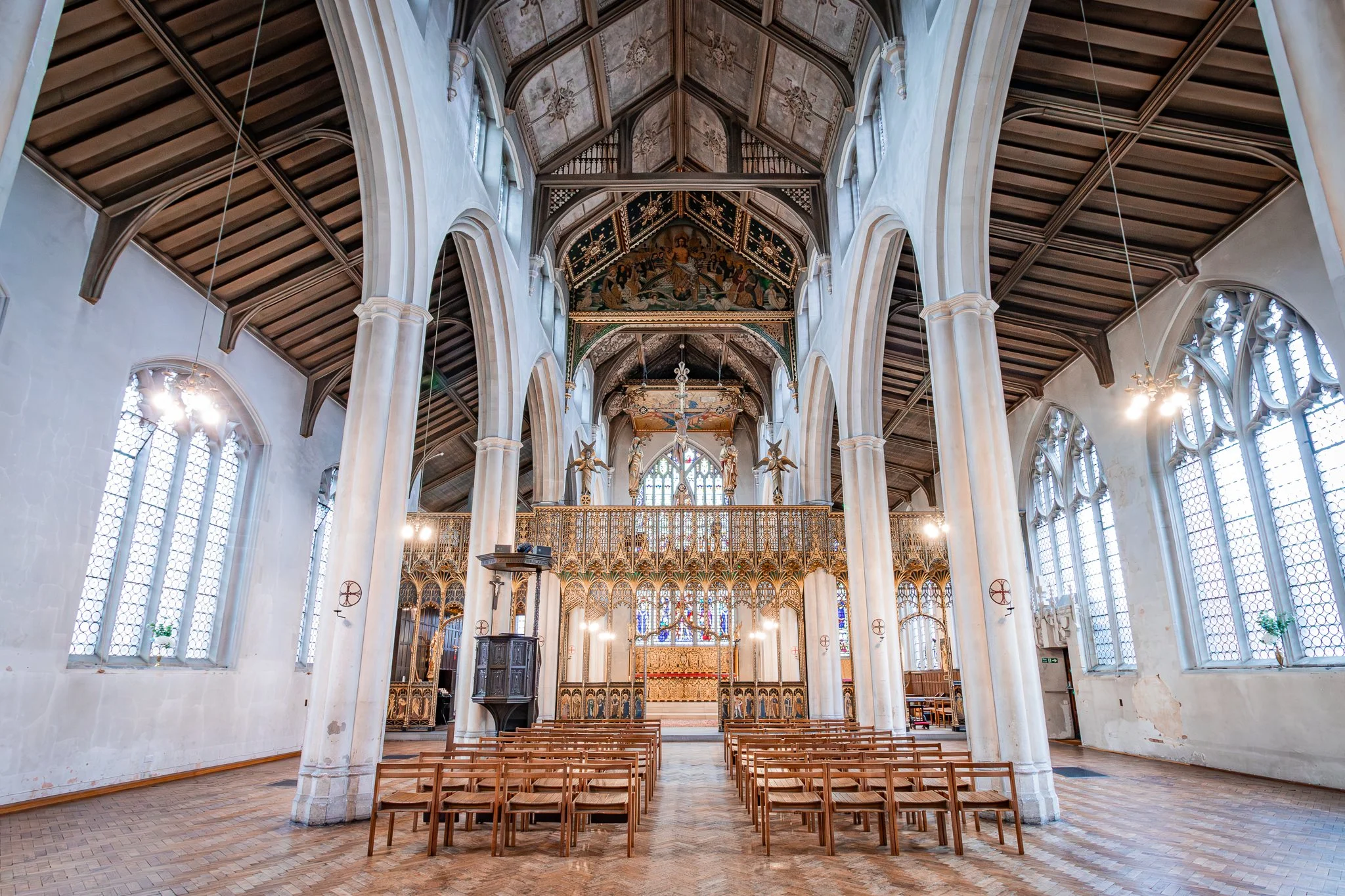 Interior of a historic church with high arched ceilings, stained glass windows, wooden pews, and ornate gold altar behind the altar.