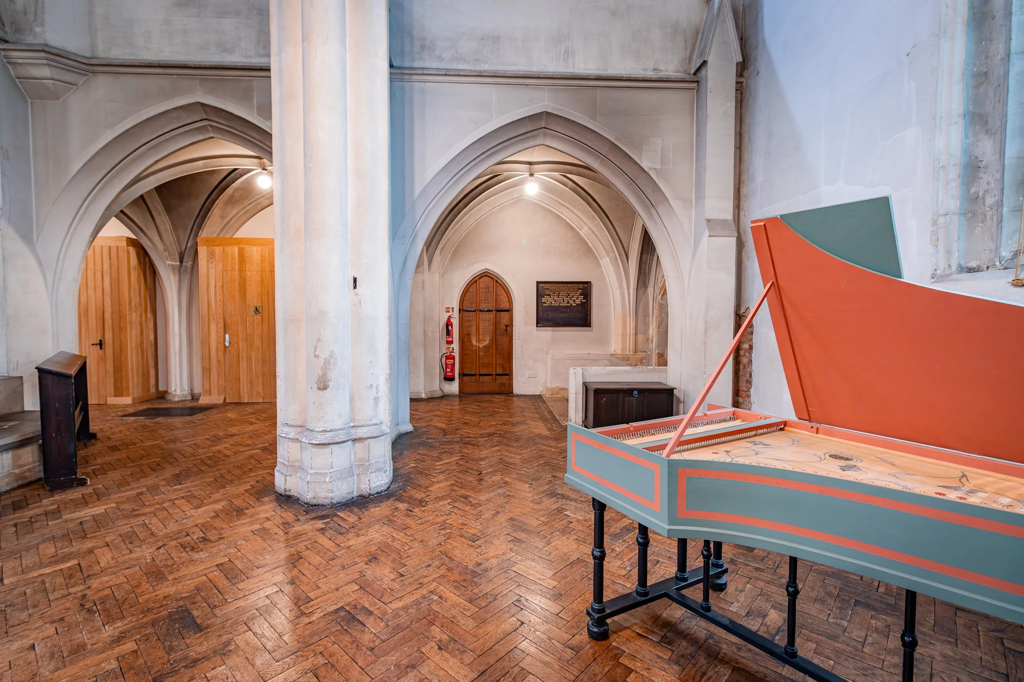 Interior of a historical building with arched ceilings, a wooden door, a vintage fire extinguisher, and a partially open pink and green piano.