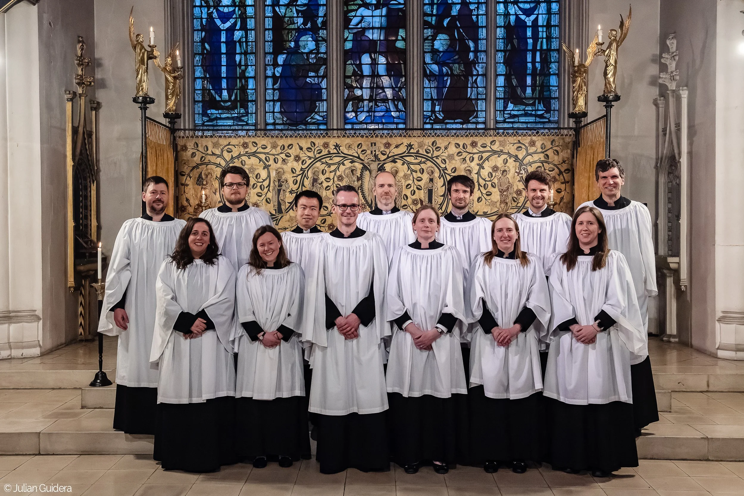 A choir of 12 people in white robes standing in a church with stained glass windows and religious artwork behind them.