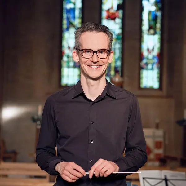 A man in a black shirt and glasses standing inside a church with stained glass windows in the background.