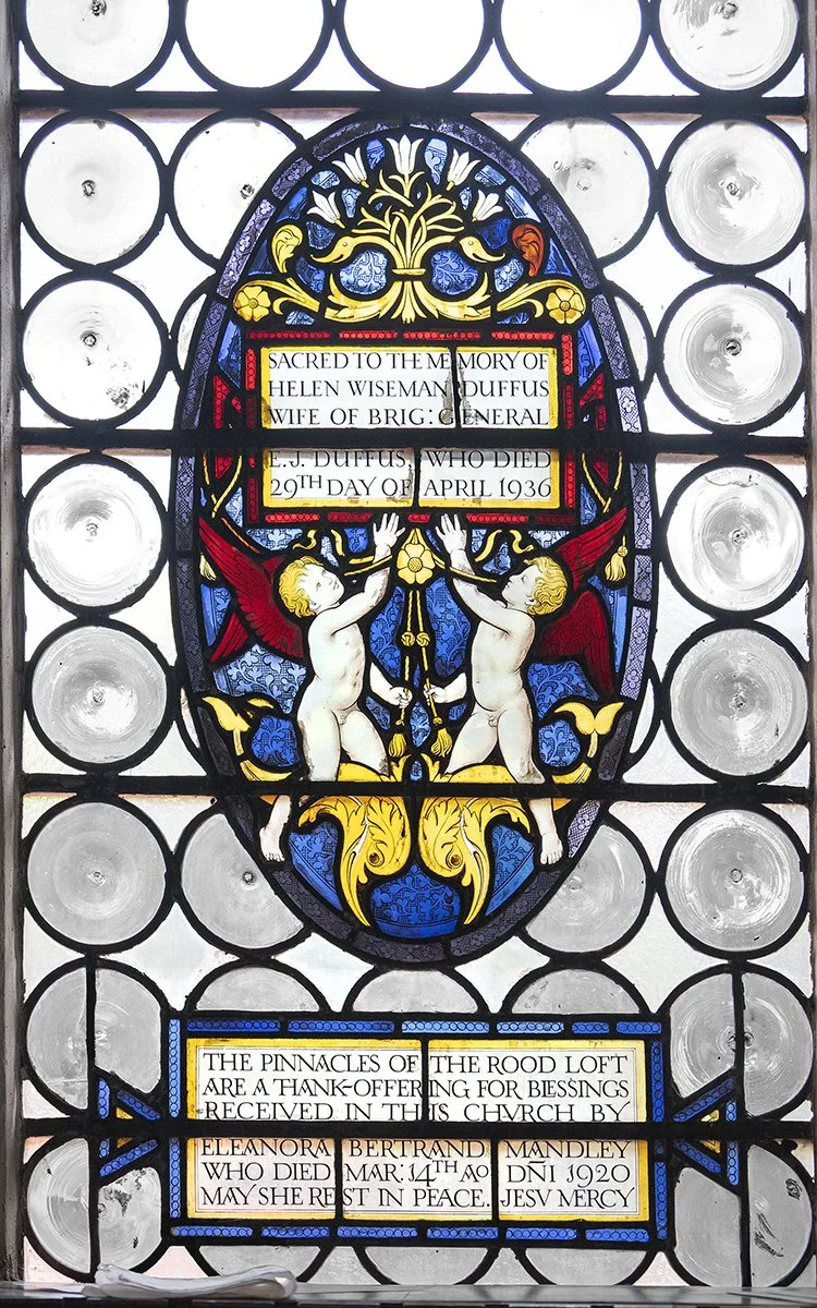 Colored stained glass window memorial with angels holding a plaque and a dedicatory inscription.