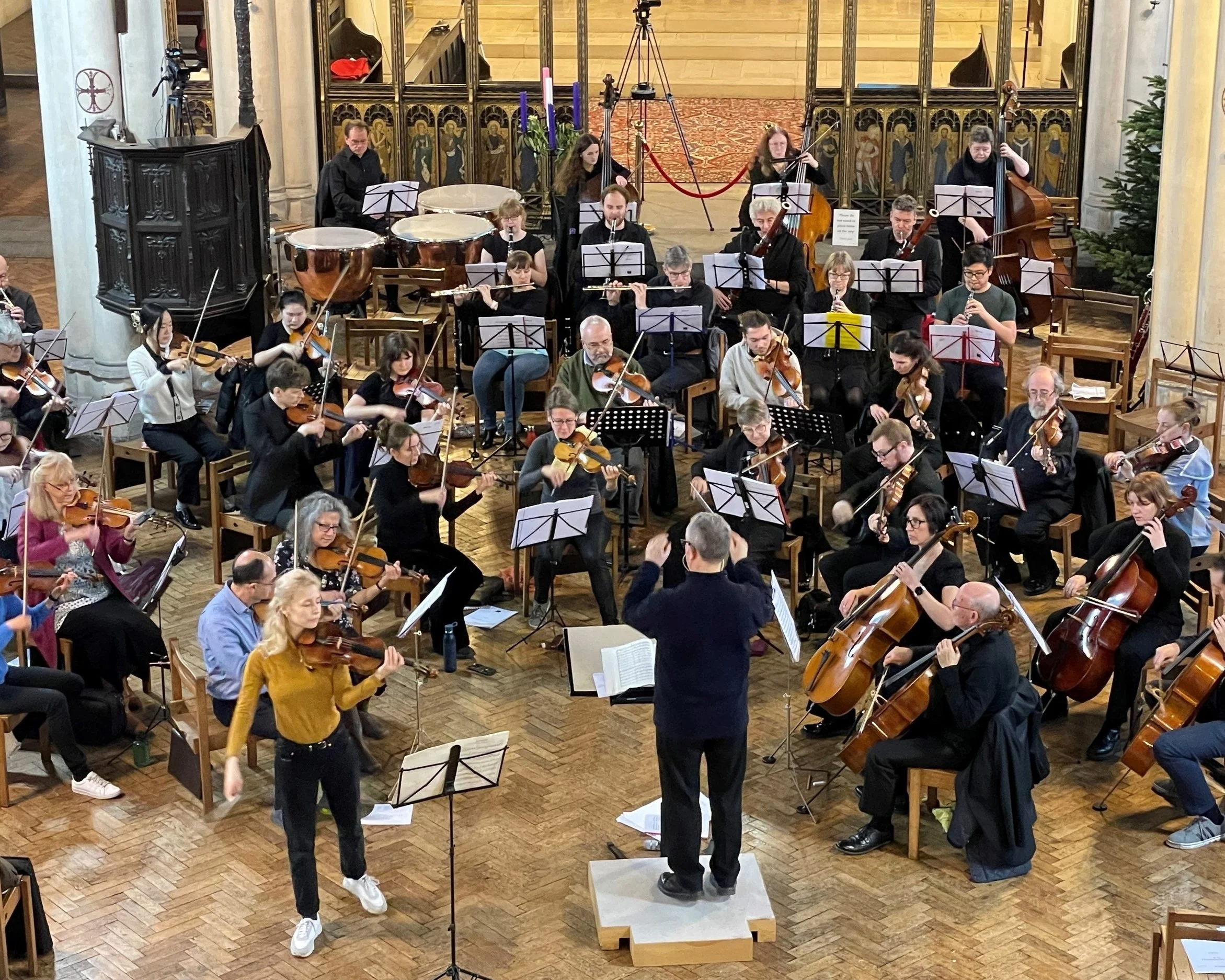 An orchestra performing in a church with a conductor, various string, wind, and percussion instruments, and an audience in the background.