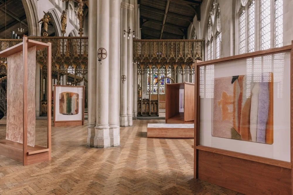 An art exhibition inside a historical church with stained glass windows, wooden framed abstract artworks on display, and tall stone columns supporting a vaulted ceiling.