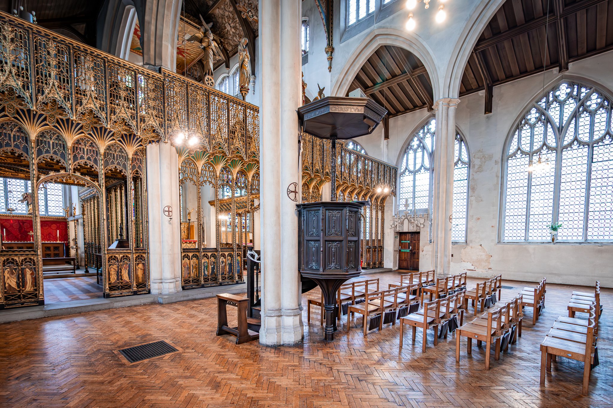 Interior of a historic church or cathedral with wooden pews, ornate gold altar railings, large Gothic windows, and a pulpit.
