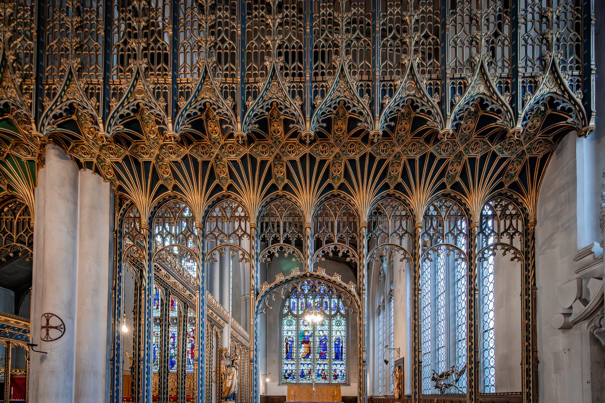 Interior of a Gothic-style church with tall stained glass windows, intricate gold and blue woodwork, statues, and a chandelier.