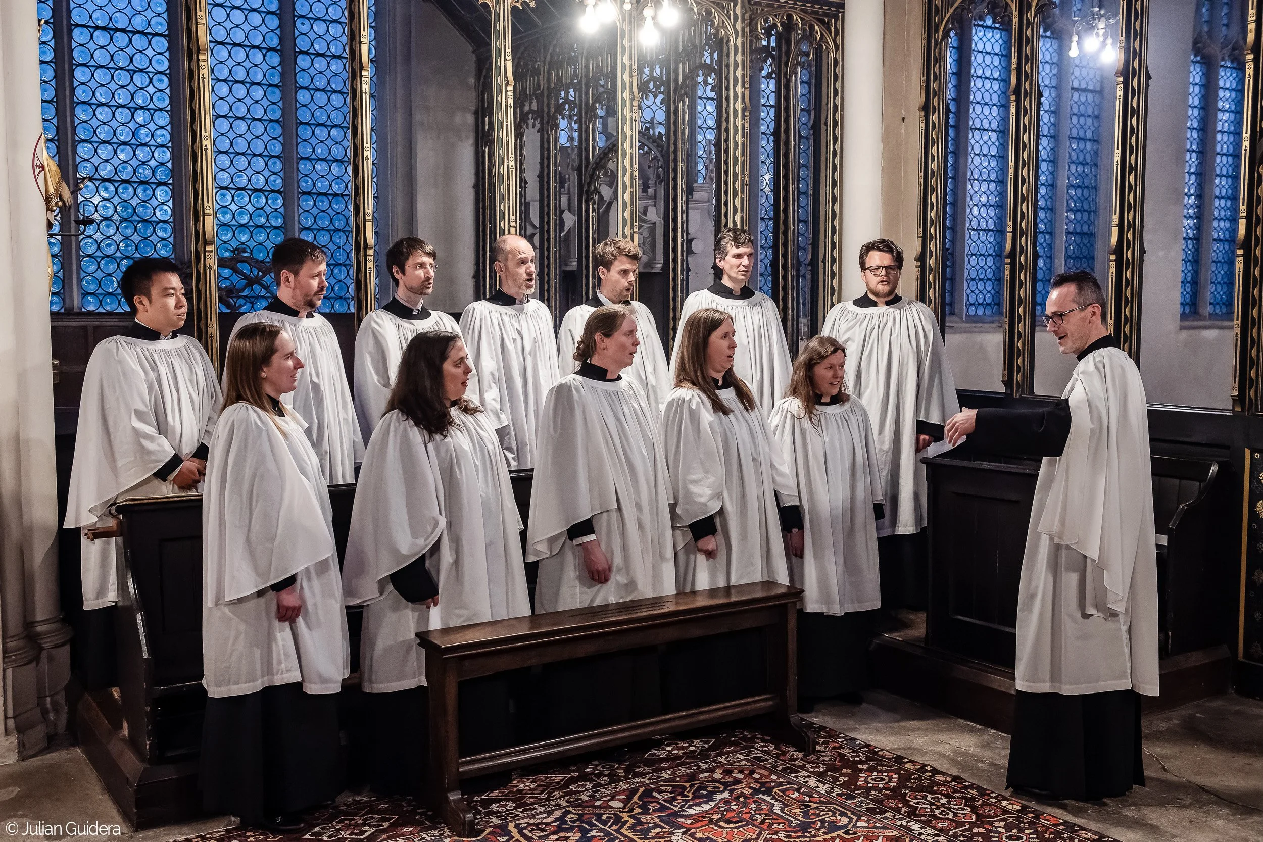 A choir of men and women dressed in white robes with black collars singing under the direction of a conductor in a church or cathedral, with stained glass windows in the background.