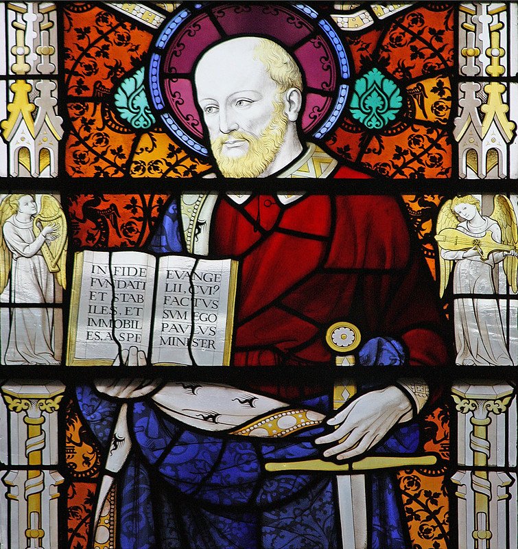 Stained glass depiction of a bearded man in a red robe holding a book, with angels and decorative patterns in the background.
