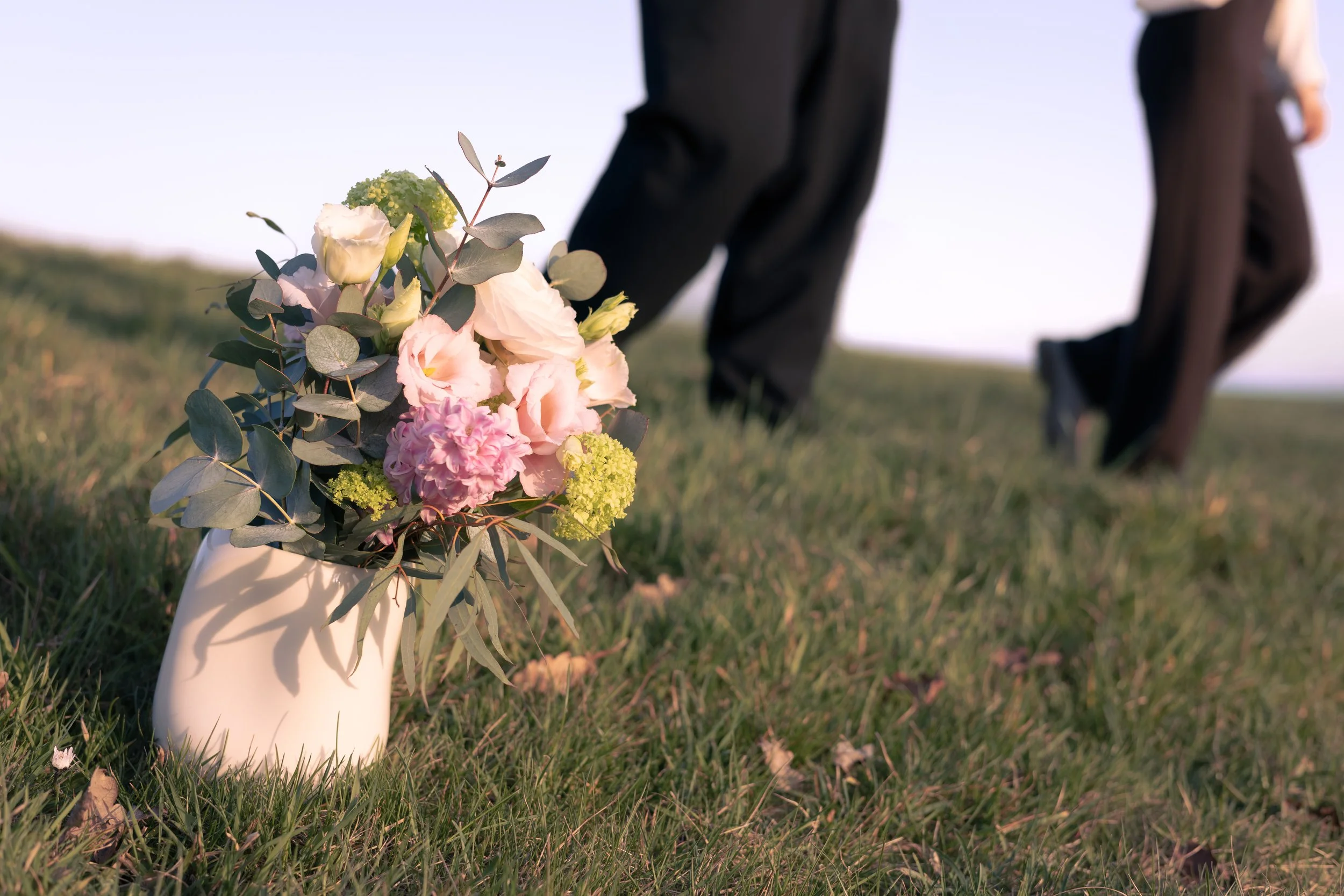 A white vase with pink and white flowers, including roses and hydrangeas, placed on a grassy field during a wedding or formal outdoor event, with two people in dark suits walking in the background.