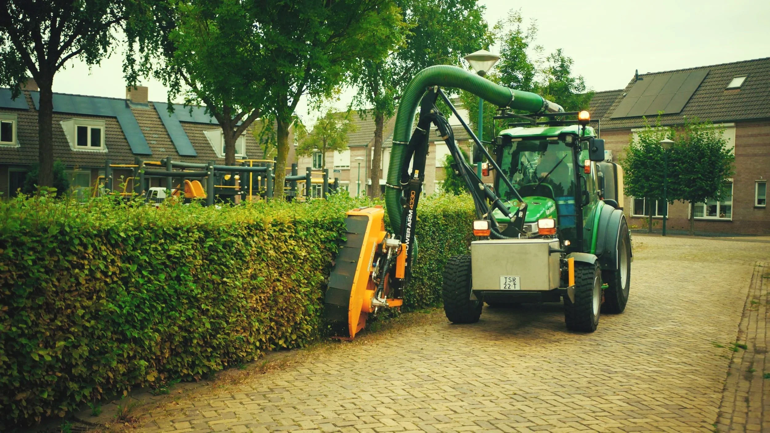 Een groene tractor met een groene uitrusting maait een heg in een buurt met woonhuizen en een speeltuin op de achtergrond.