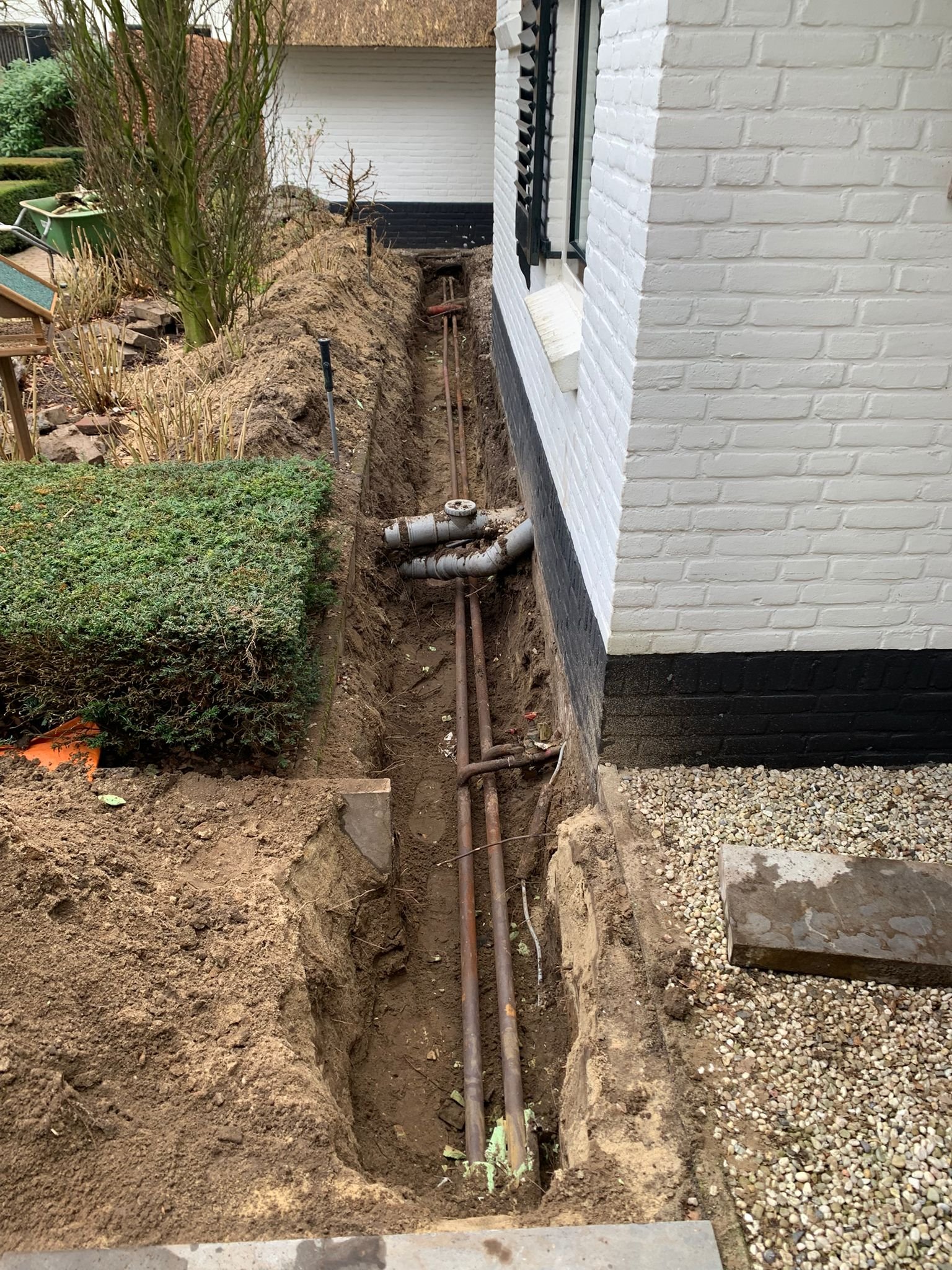 Bouwput met PVC-buizen en aardewerk pijpleidingen naast een witte gevel van een huis.