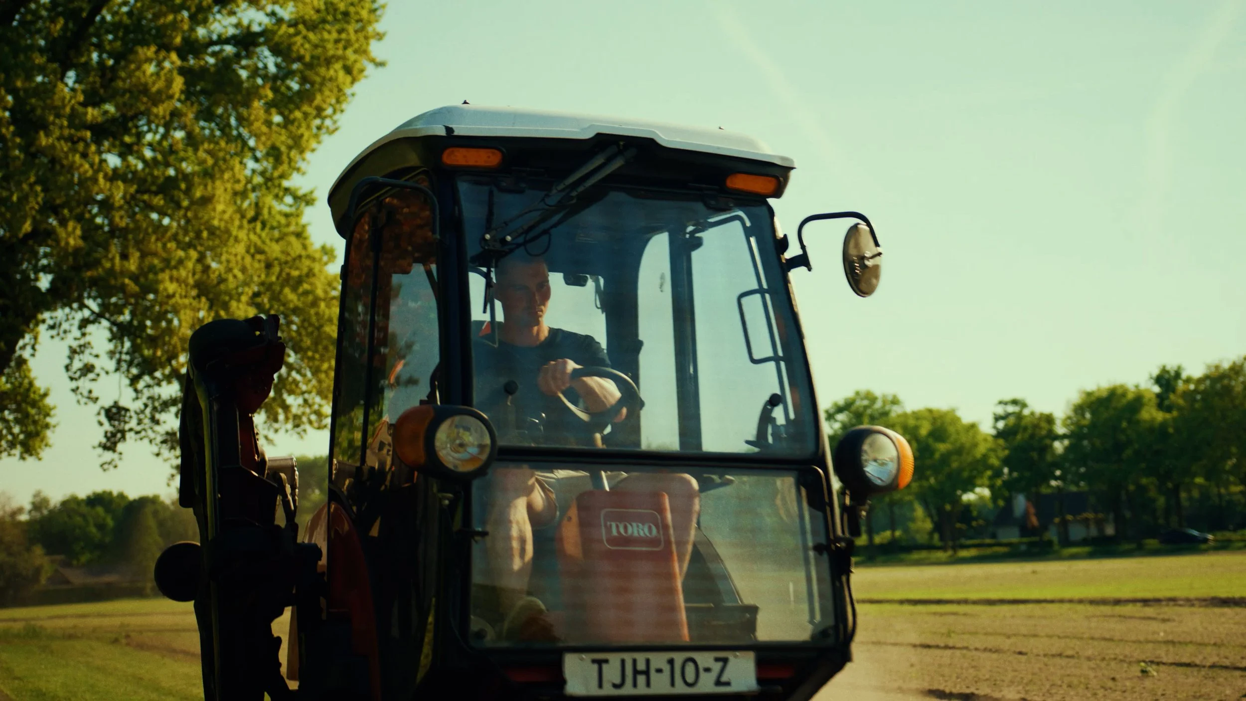 Man rijdt een tractor op een grasveld met bomen op de achtergrond, onder een heldere lucht.