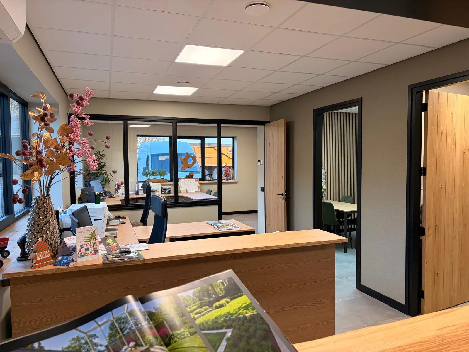 Kantoorruimte met bureau, computer, bloemen en brochures, met glazen scheidingswanden en een witte plafond met ingebouwde verlichting.