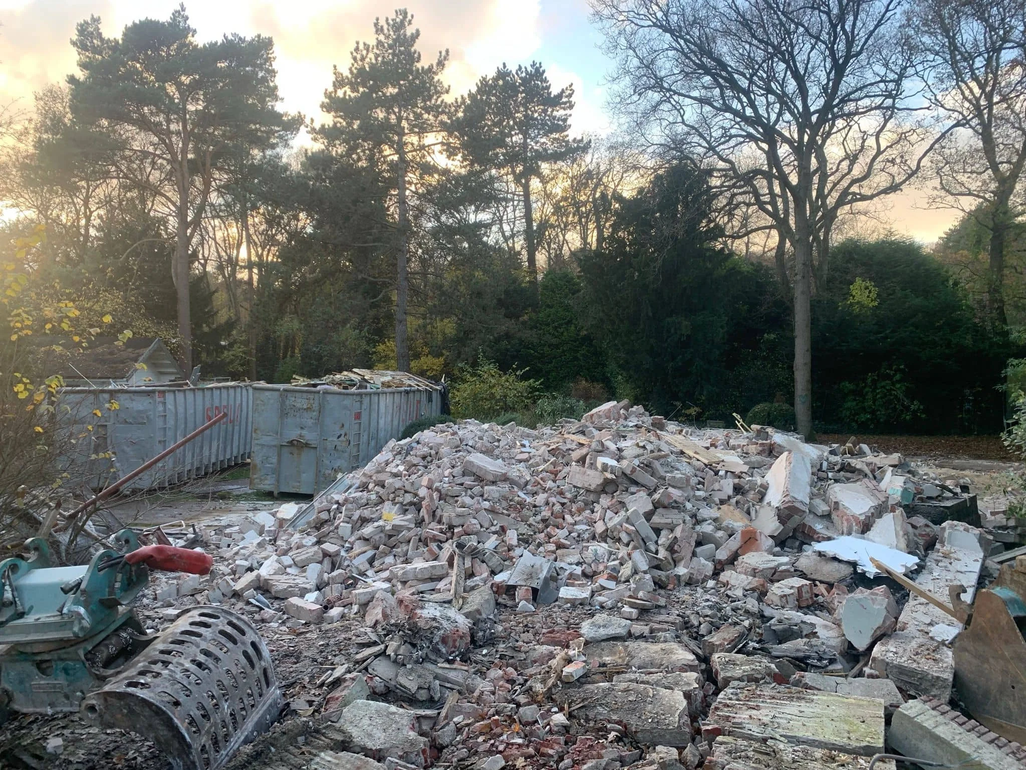 Een bouwplaats met een grote hoop stenen en puin, een container, en een tuin met bomen op de achtergrond. Het lijkt op sloopwerk of renovatie.