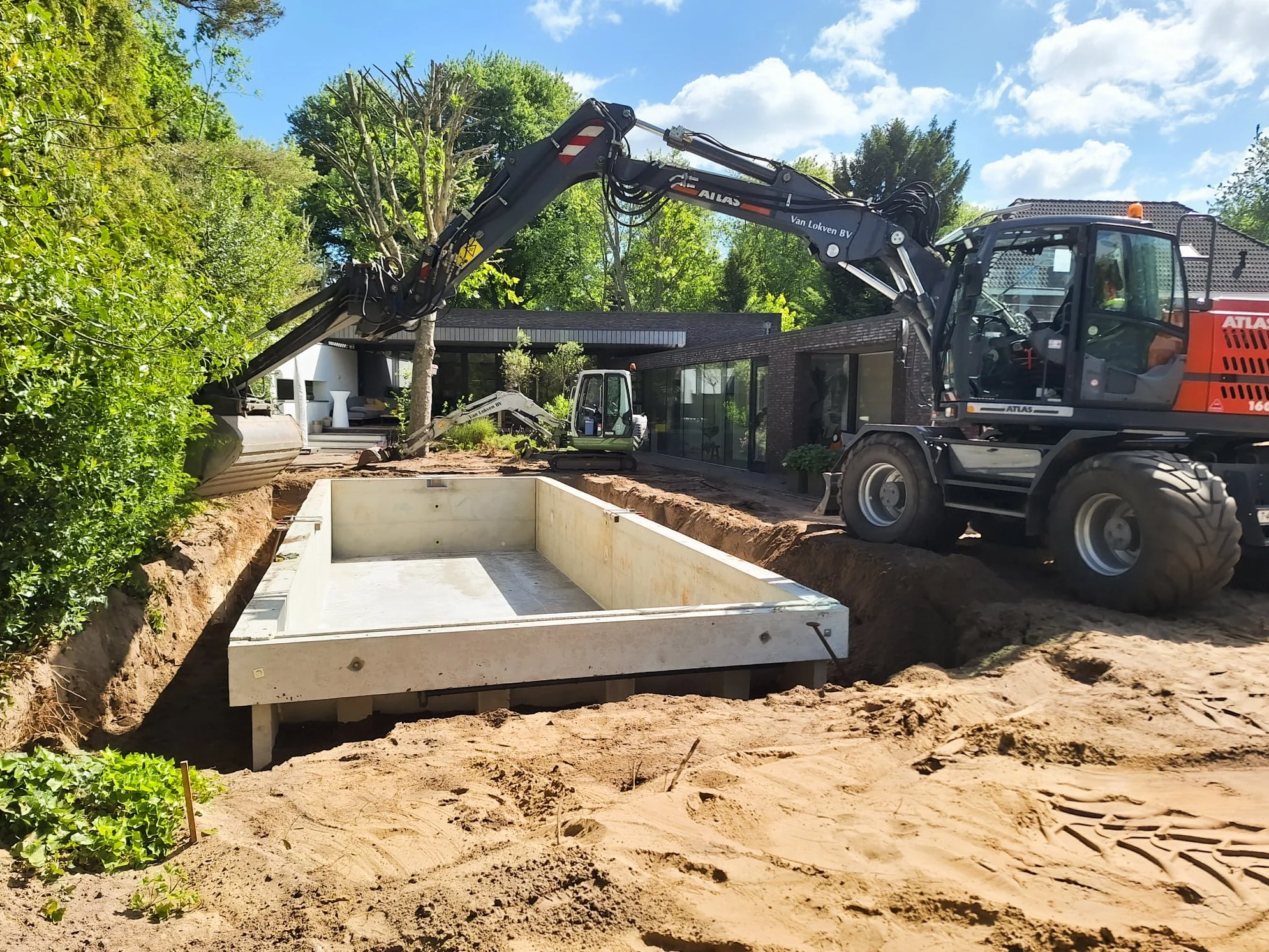 Een bouwplaats met graafmachines en een uitgegraven fundering voor een nieuw bouwwerk, naast een modern huis met grote ramen en een tuin. voor het ingraven van een zwembad
