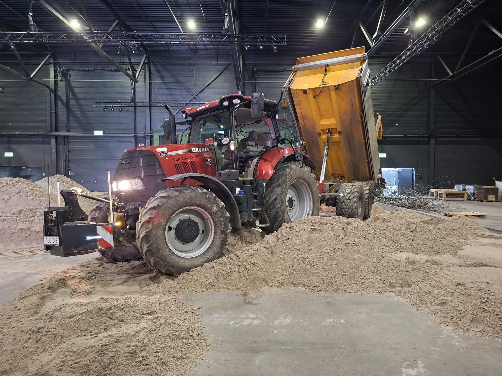 Een rode tractor in een binnenruimte die zand of grind verplaatst, naast een grote kiepwagen gevuld met zand.