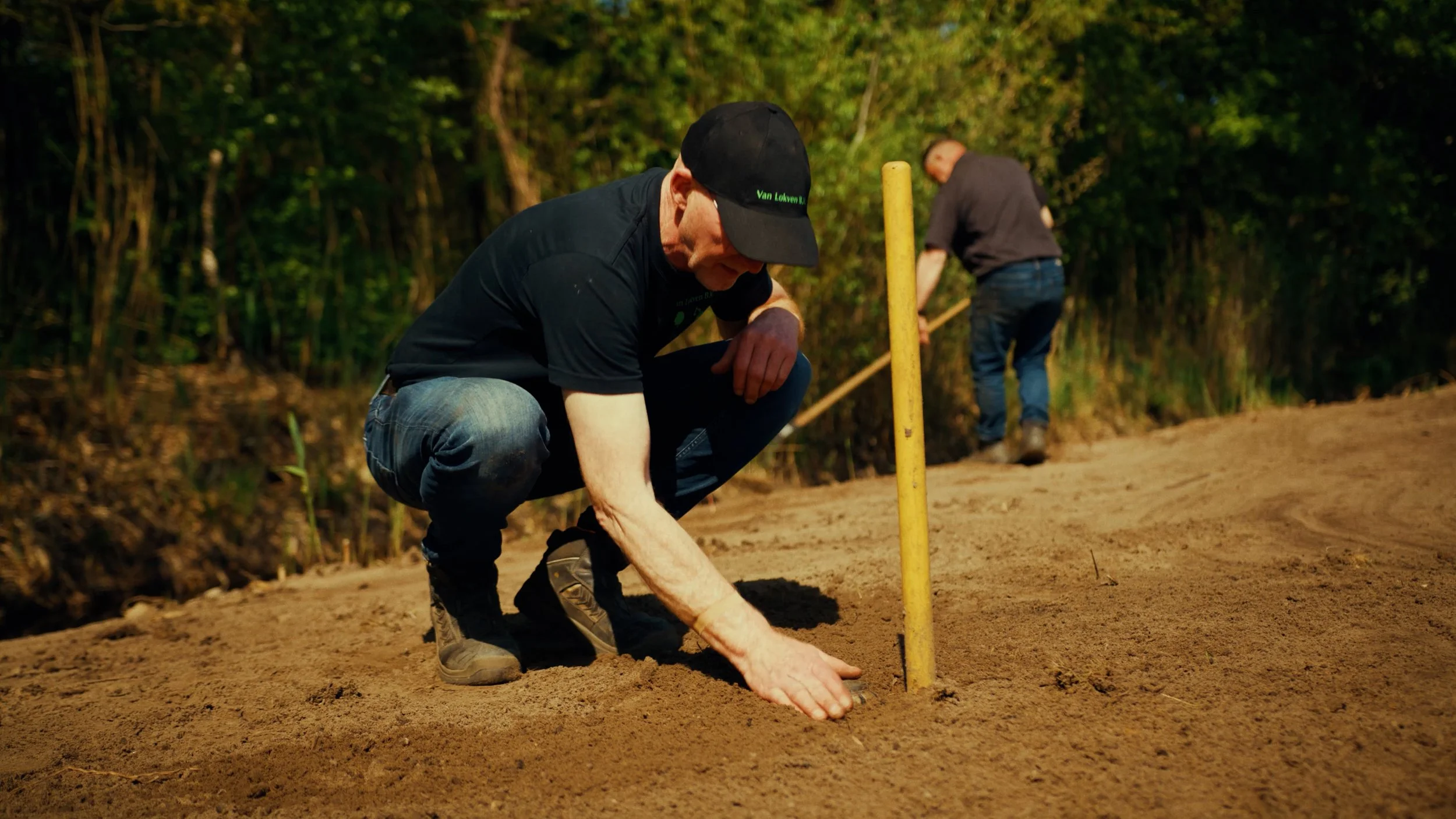 Twee mannen werken op een golfbaan, met groene bomen op de achtergrond, een ervan is aan het graven of planten en de ander staat verder weg.