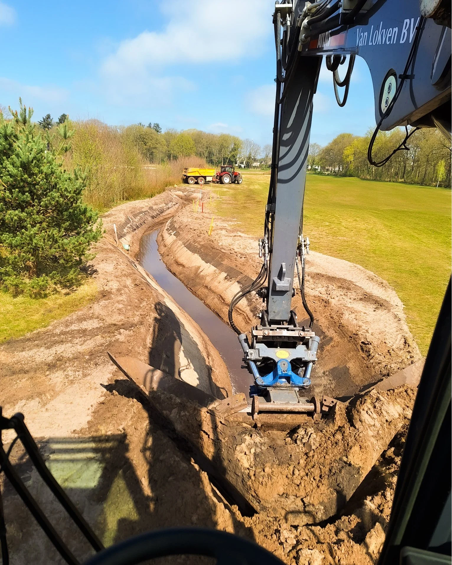Het beeld toont een bouwplaats met een graafmachine die een geul graaft voor een waterleiding of pijpleiding. In de achtergrond is een tractor met een trailer te zien, terwijl het landschap bestaat uit grasvelden en bomen onder een blauwe hemel met enkele wolken.