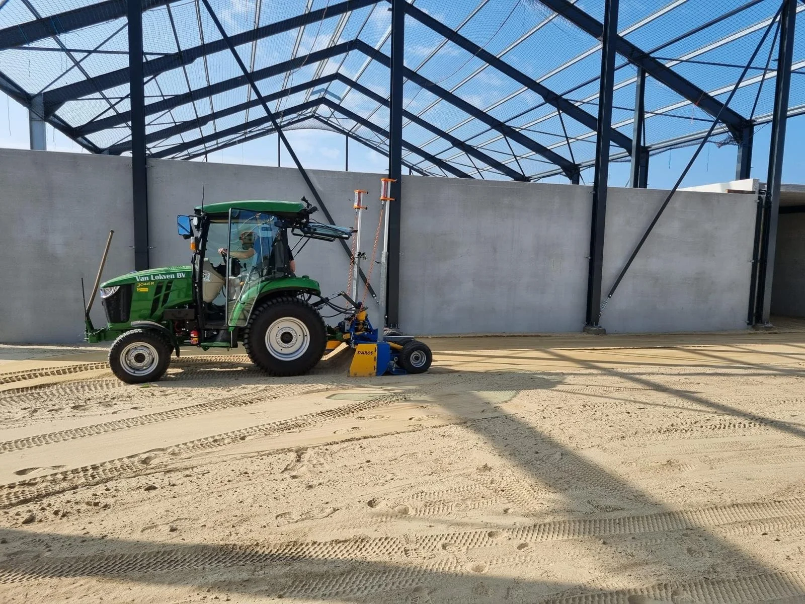 Een groene tractor die de vloer van een gebouw afwerkt met een blauwe machine, onder een grote glazen dak en een betonnen muur. laser kilverbord