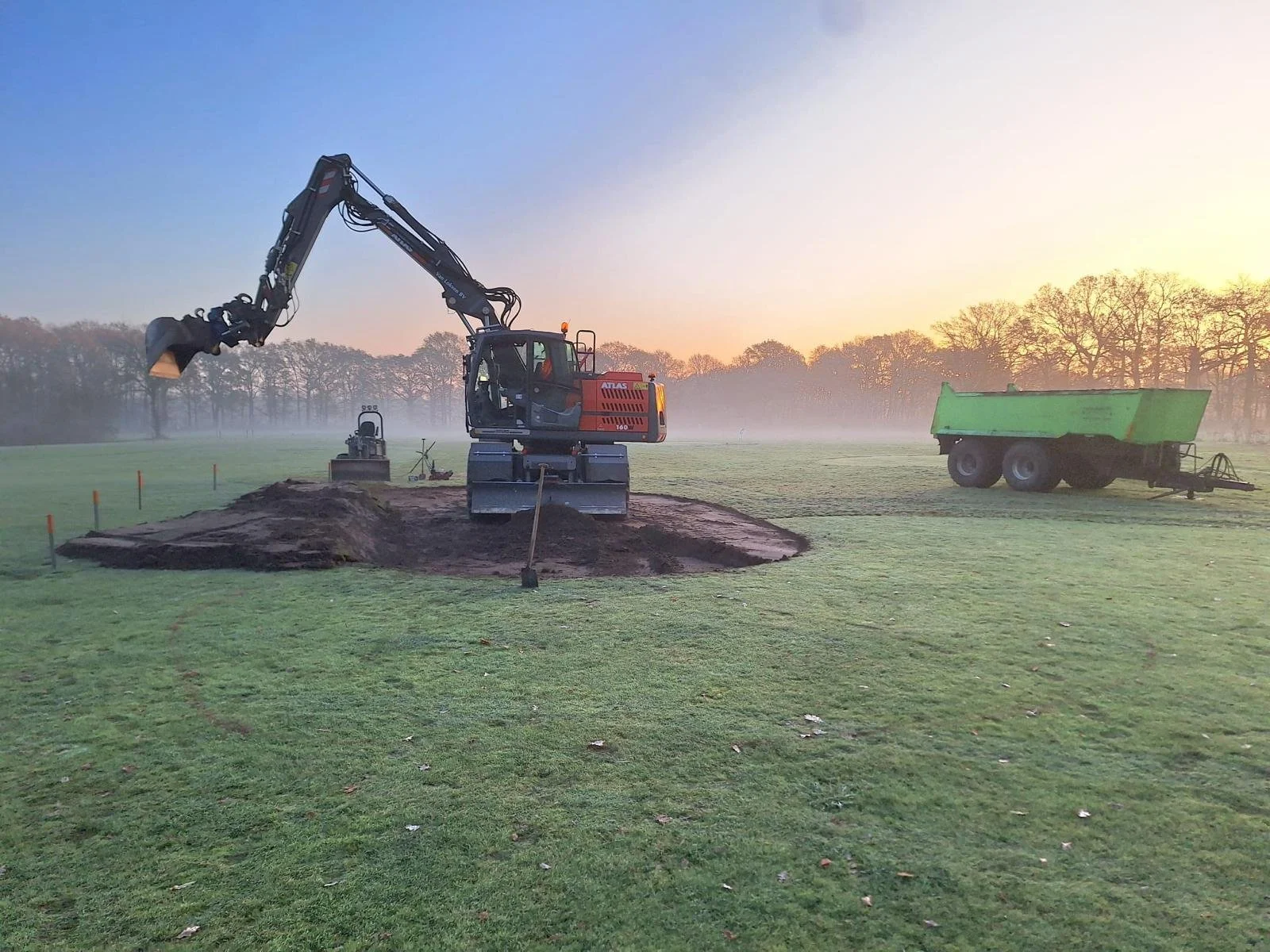 Een graafmachine die op een grasveld een gat graaft, met een trailer ernaast, in de ochtend met een mistige achtergrond en bomen in de verte.
