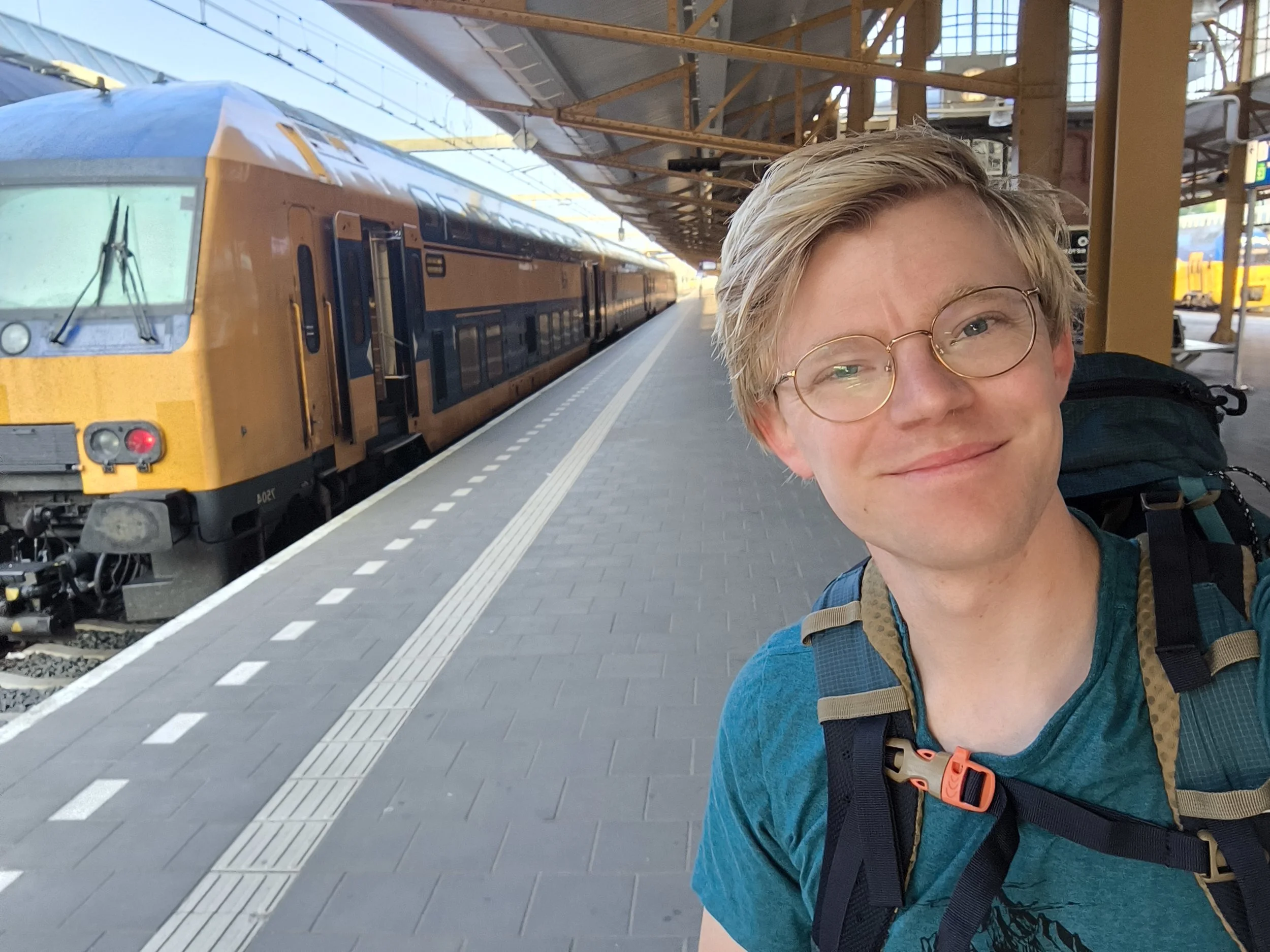 Lachende jongen met rugzak op een station, met een geel en blauw trein op de achtergrond.