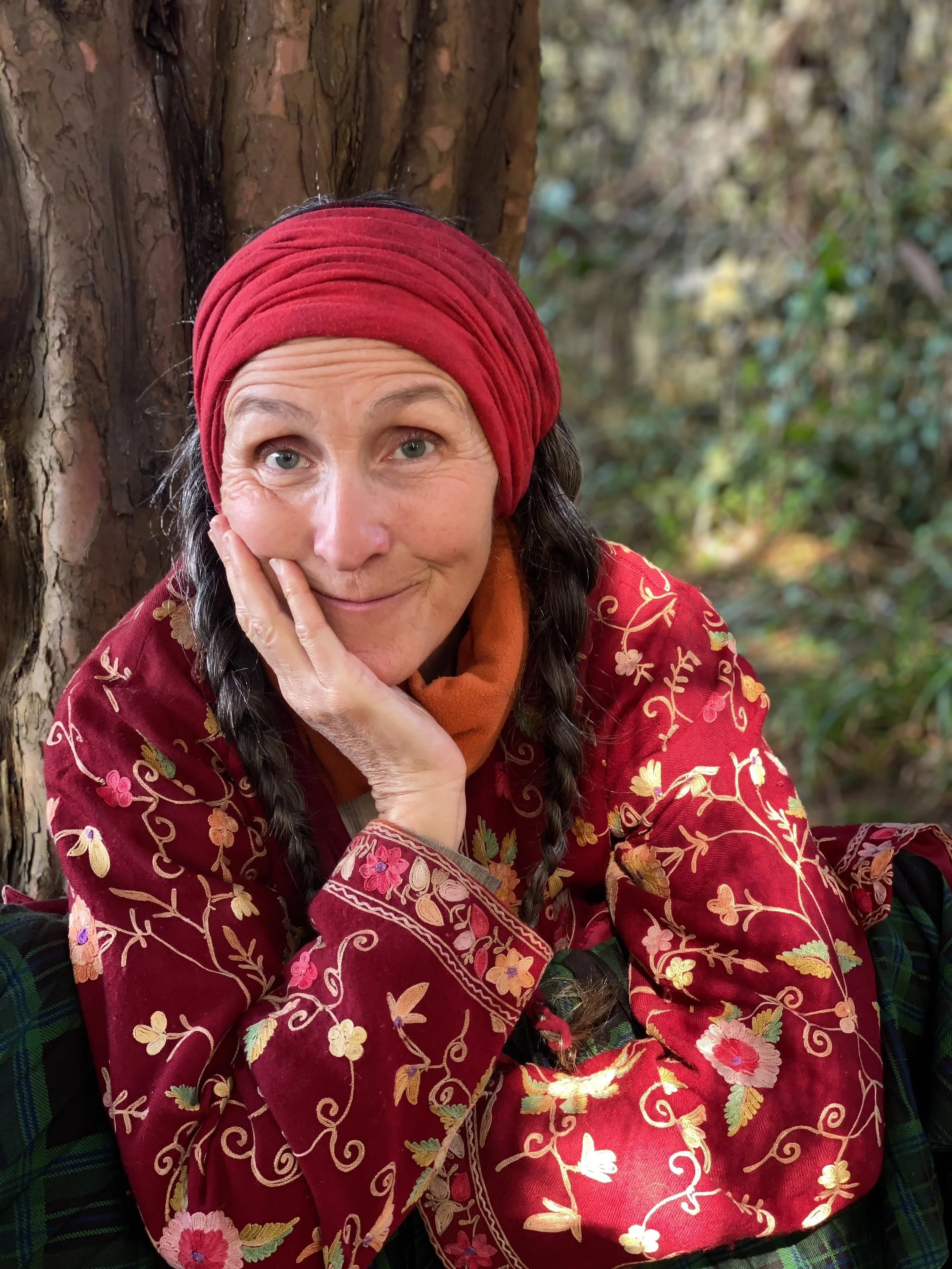 A woman with gray hair and blue eyes, wearing a red headscarf and a red embroidered jacket, sitting outdoors next to a tree with a forest background.
