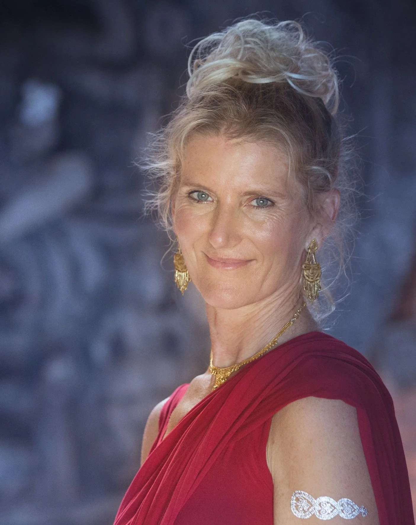 Shiva Rea yoga teacher dressed in red wearing golden earrings and necklace smiling