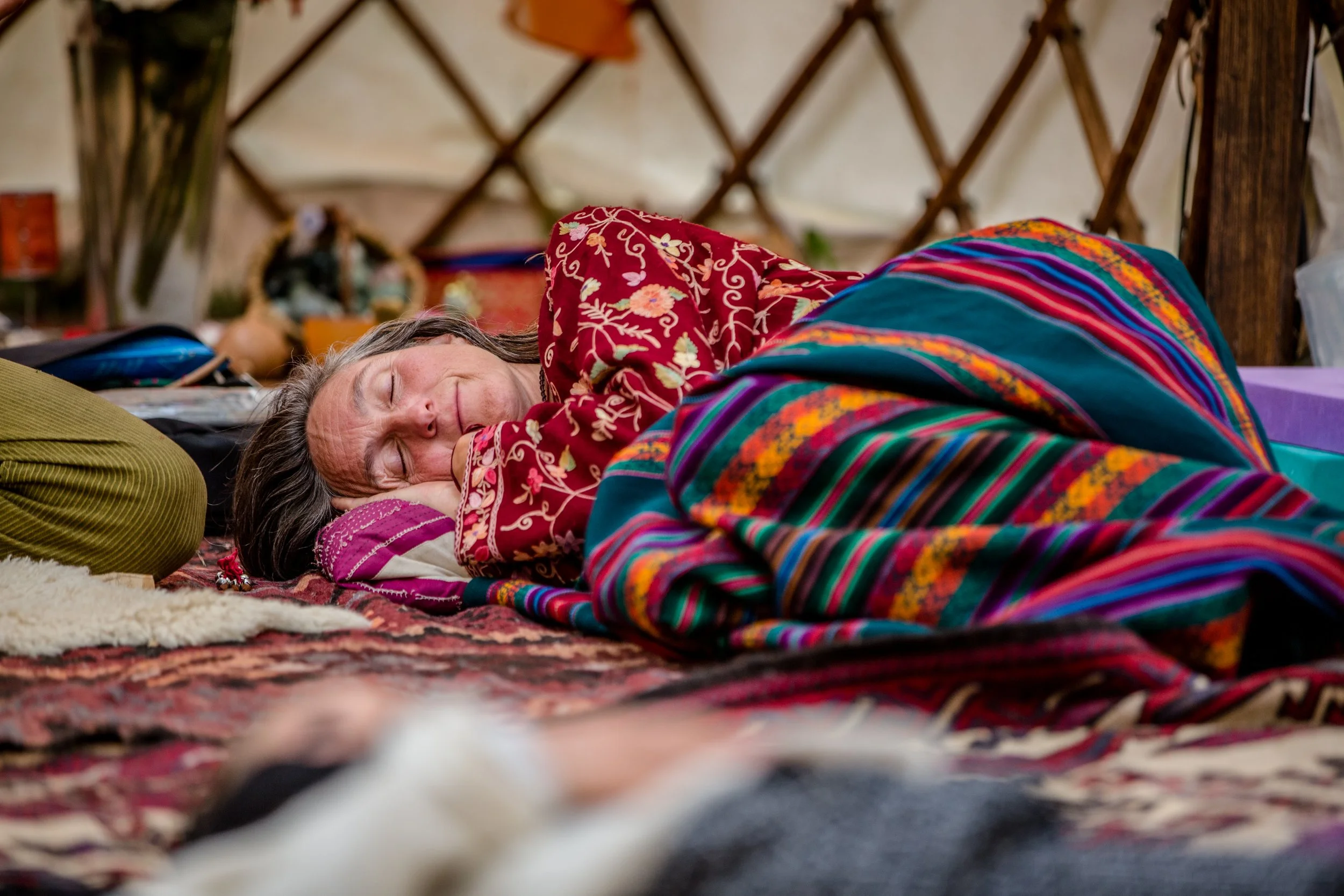A woman with long gray hair, wearing a red embroidered shirt, sleeping on a colorful patterned blanket in a cozy, rustic setting.