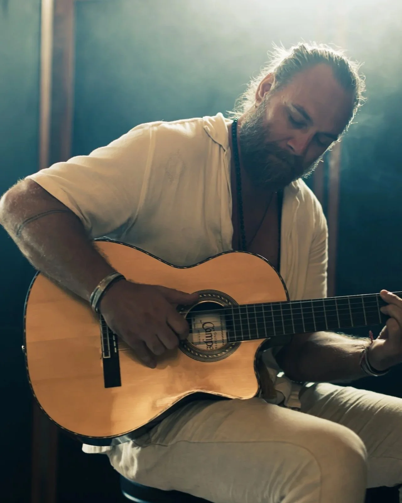 A man with a beard and long hair tied back playing an acoustic guitar in a dimly lit room.
