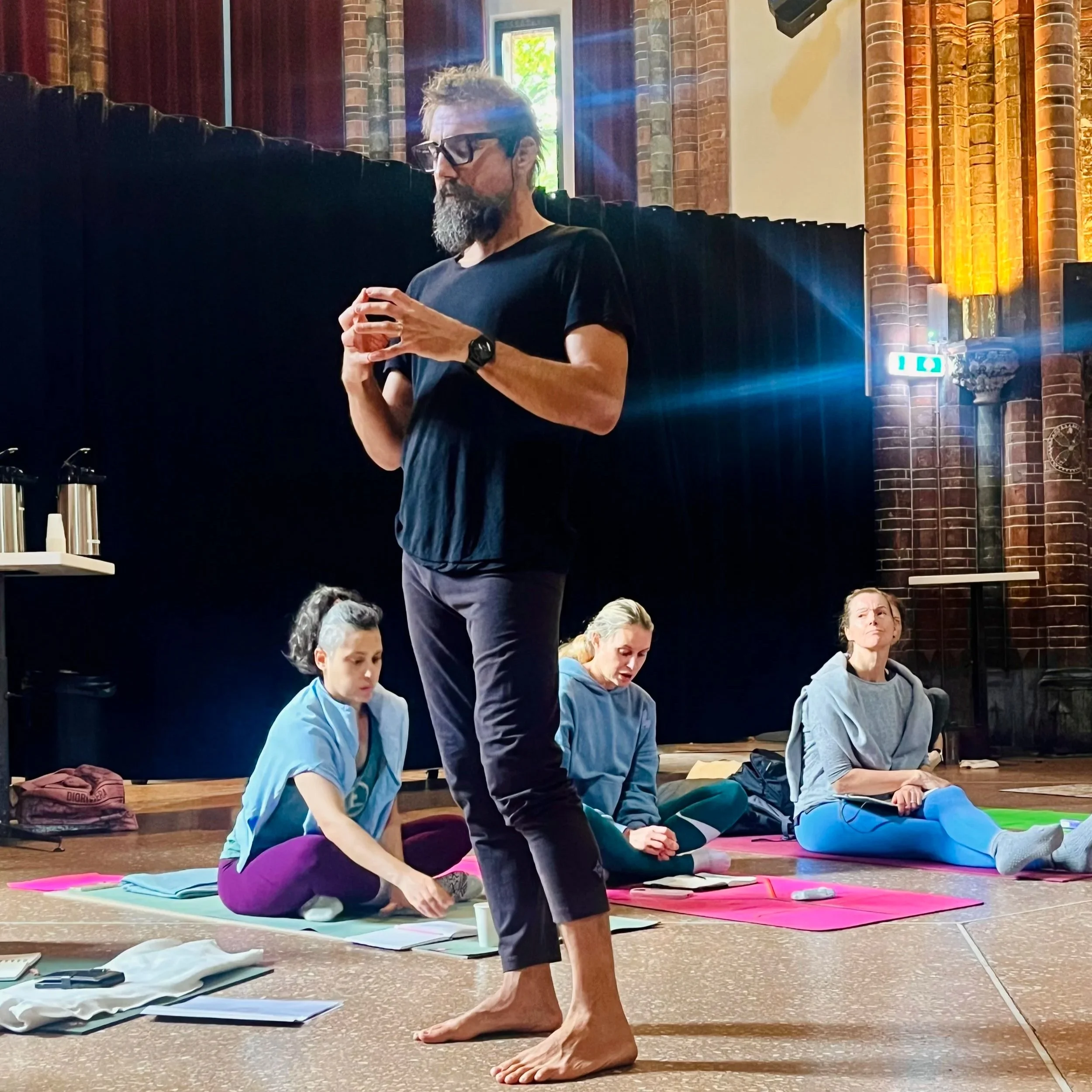A yoga instructor stands barefoot in front of three women practicing yoga or meditation, seated on yoga mats in a spacious room with brick walls and high windows, during daytime.