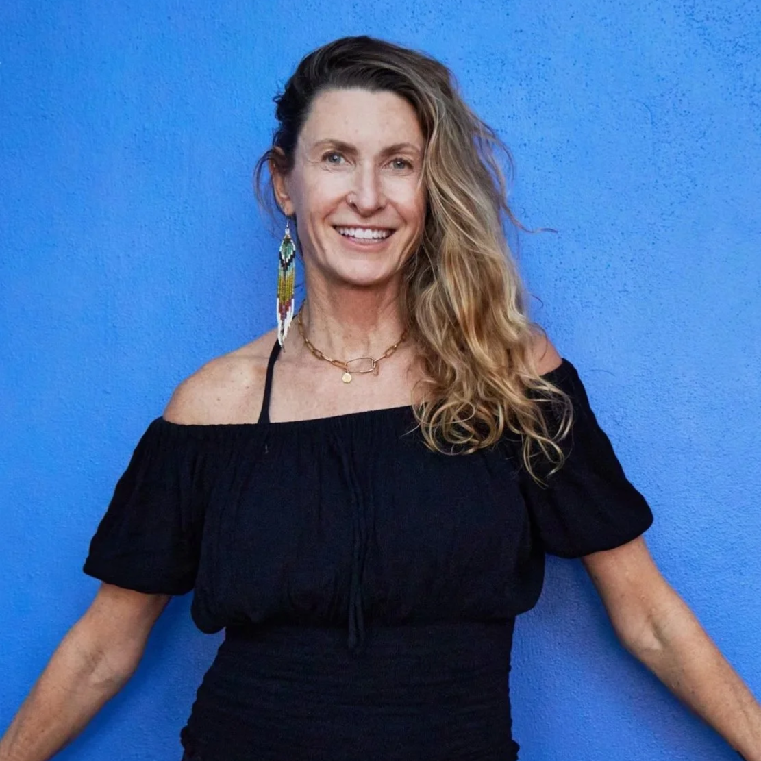 A woman with wavy blonde hair wearing a black off-the-shoulder top, colorful beaded earrings, and layered necklaces, standing against a vibrant blue wall and smiling.
