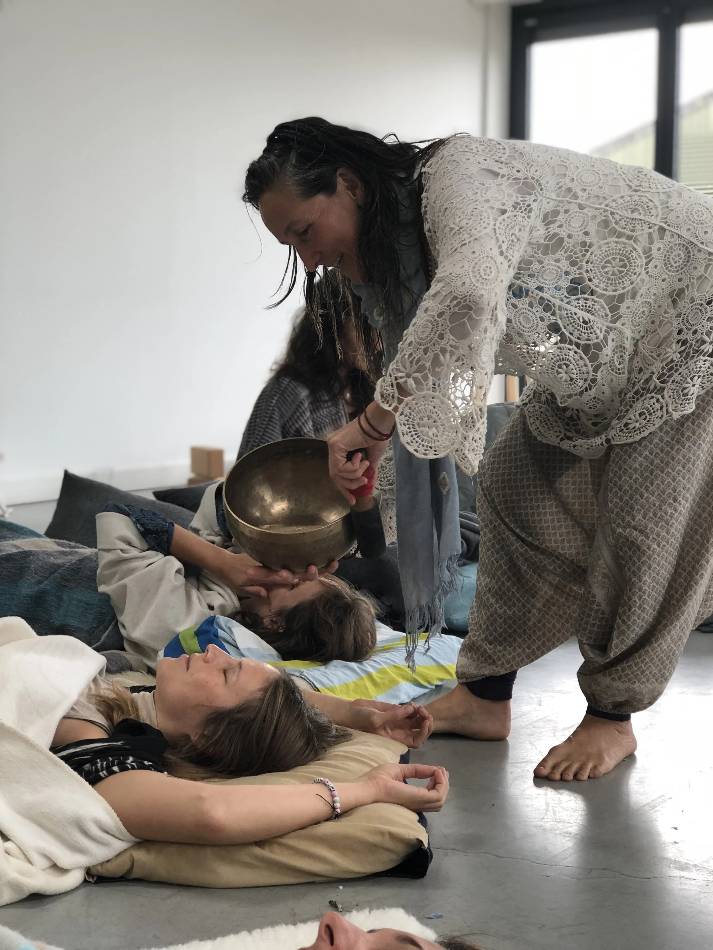 Women participating in a yoga or meditation session on the floor, with one woman using a singing bowl and others lying down, in a bright room with large windows.