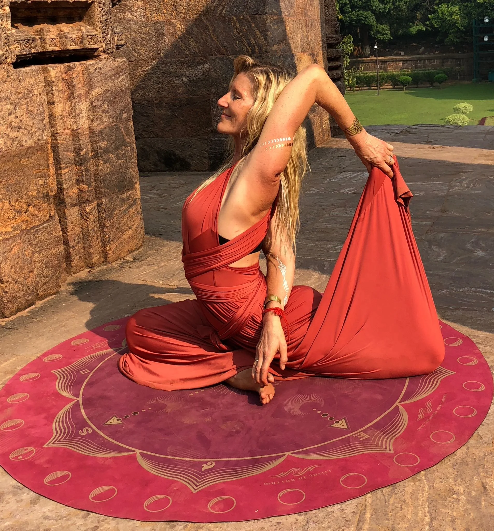 A woman practicing yoga outdoors on a red circular yoga mat, wearing a red dress, sitting in a seated backbend pose with one arm reaching behind her head, holding her foot, in front of a stone structure and green landscape.