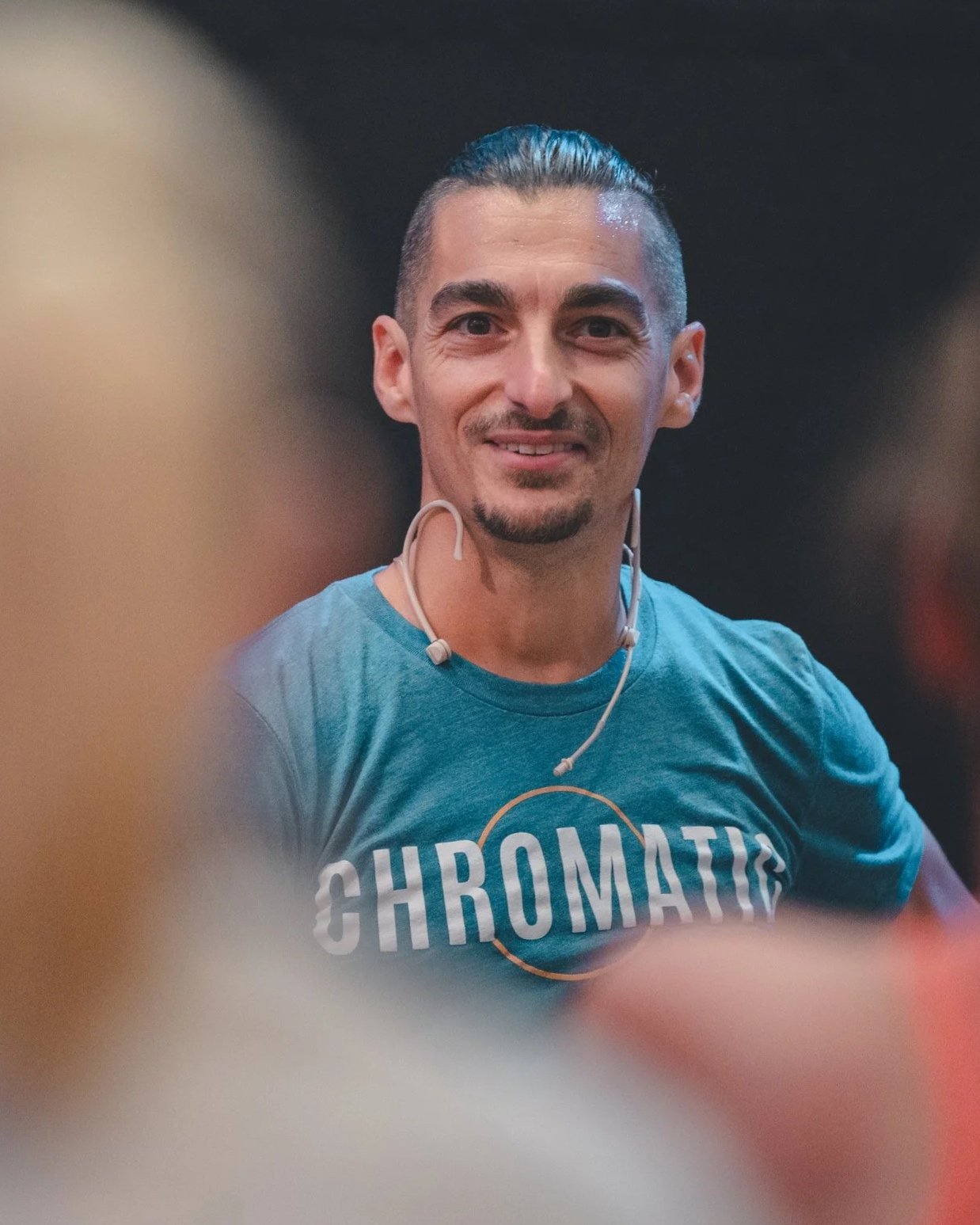 A man with slicked-back hair, wearing a blue T-shirt with the word 'CHROMATIC' on it, is smiling and wearing a headset.
