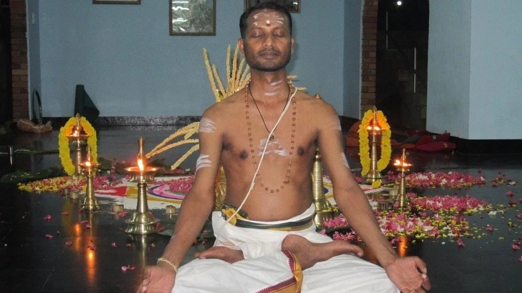 A man dressed in traditional Indian attire participating in a spiritual ritual with oil lamps, flower petals, and decorative elements around him.