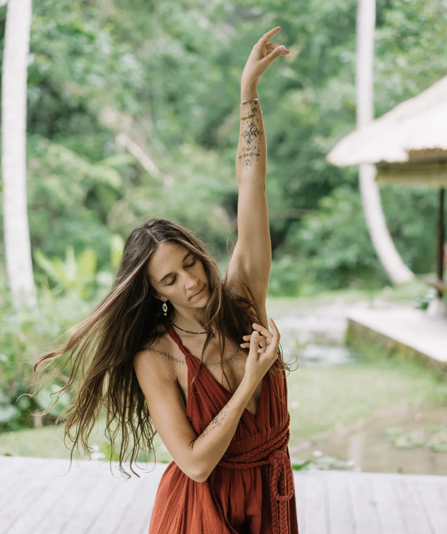 A woman in a red dress with bohemian tattoos on her arm and neck, standing outdoors in a lush green environment, raising her arm in a graceful pose with her eyes closed.