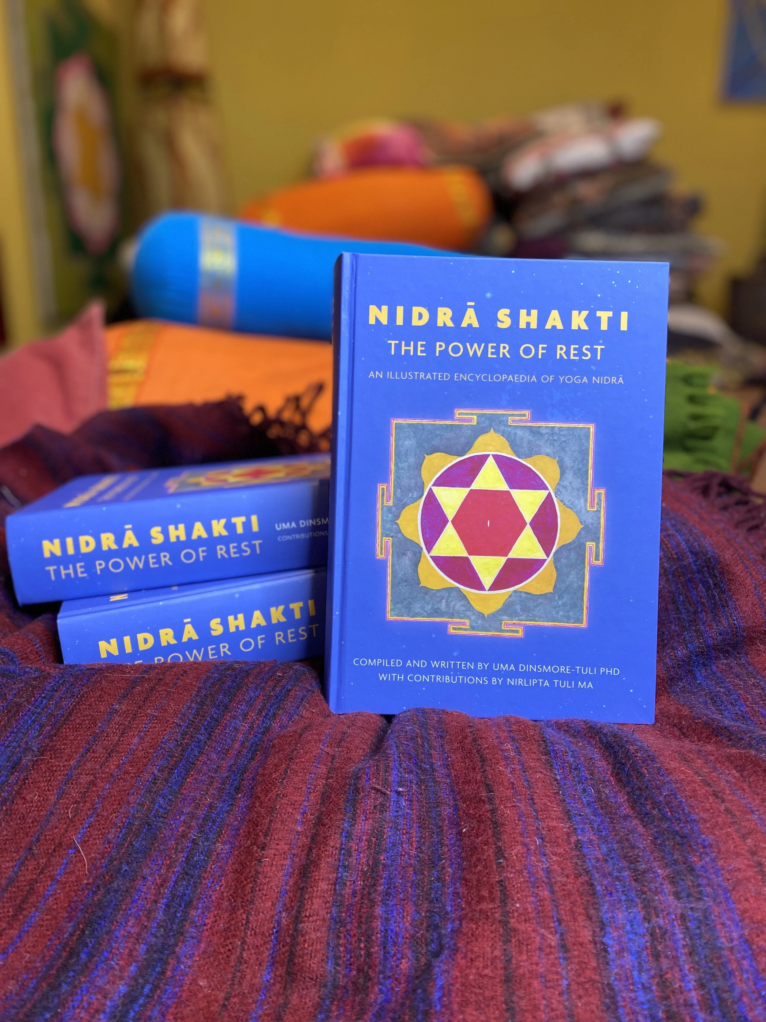 Blue book titled 'Nidra Shakti: The Power of Rest' standing upright with similar books lying behind it on a multicolored textile surface.