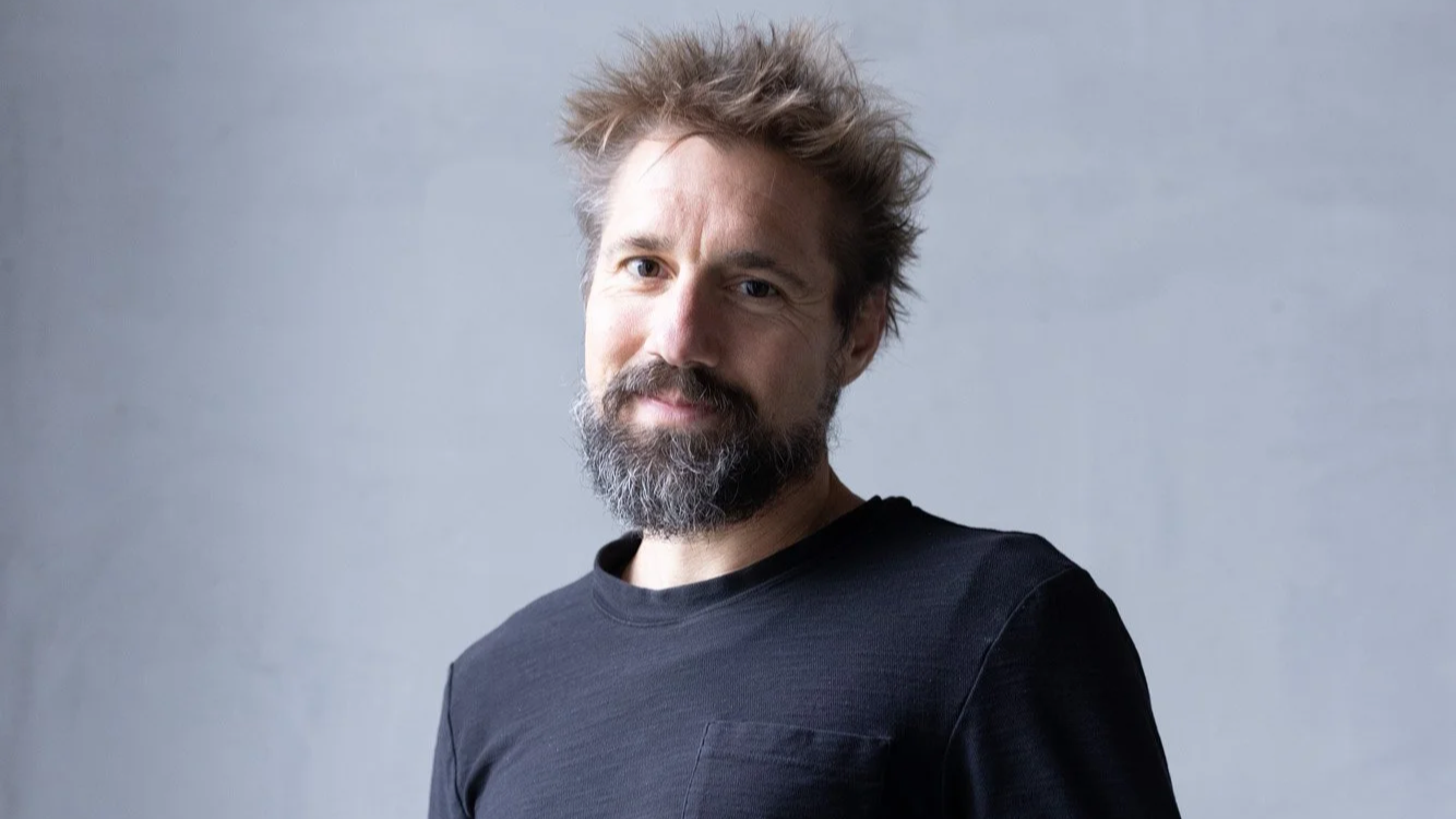 A man with a beard and tousled hair wearing a black shirt, looking at the camera against a plain gray background.