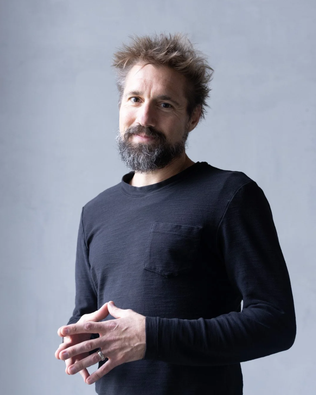 A man with a beard and tousled hair wearing a black long-sleeve shirt, standing against a plain gray background. He is smiling slightly and clasping his hands in front of him.