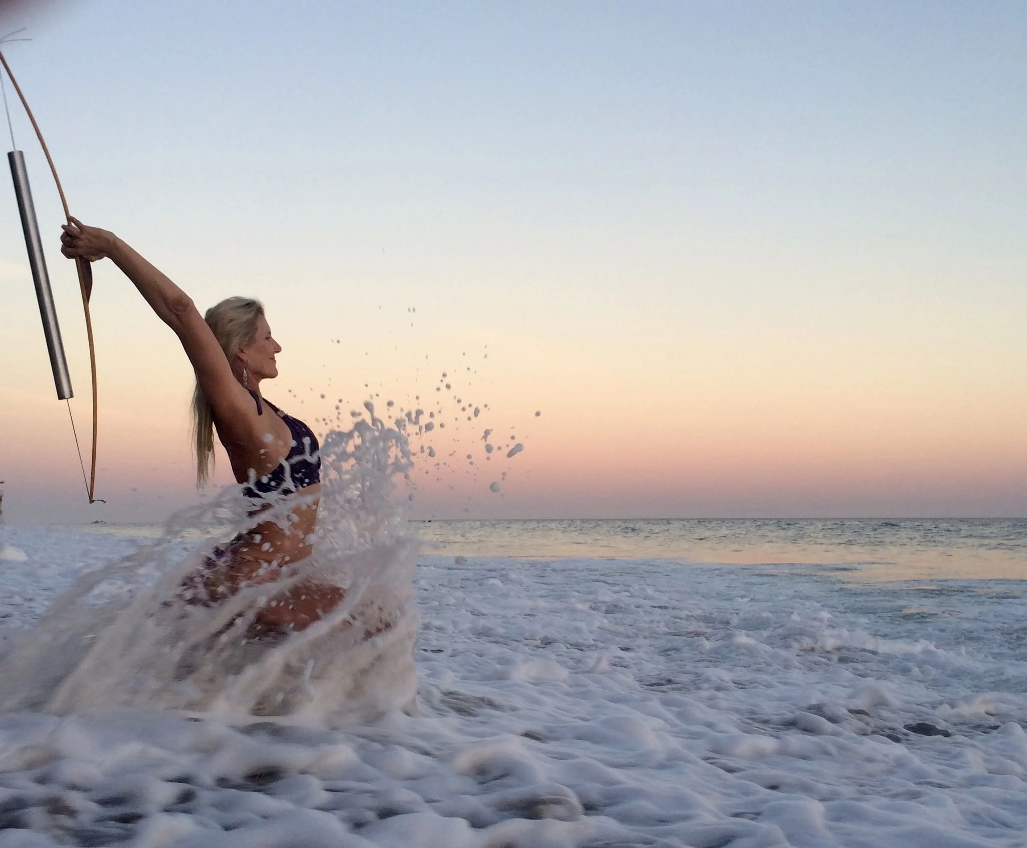 A woman in a bikini holding a bow and arrow on a beach at sunset, with waves splashing around her and colorful sky in the background.