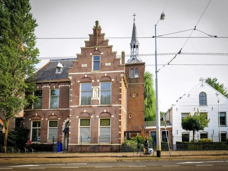 A historic red brick building with ornate architectural details and a small church steeple, next to a white building with a tree and a person riding a bicycle in front.