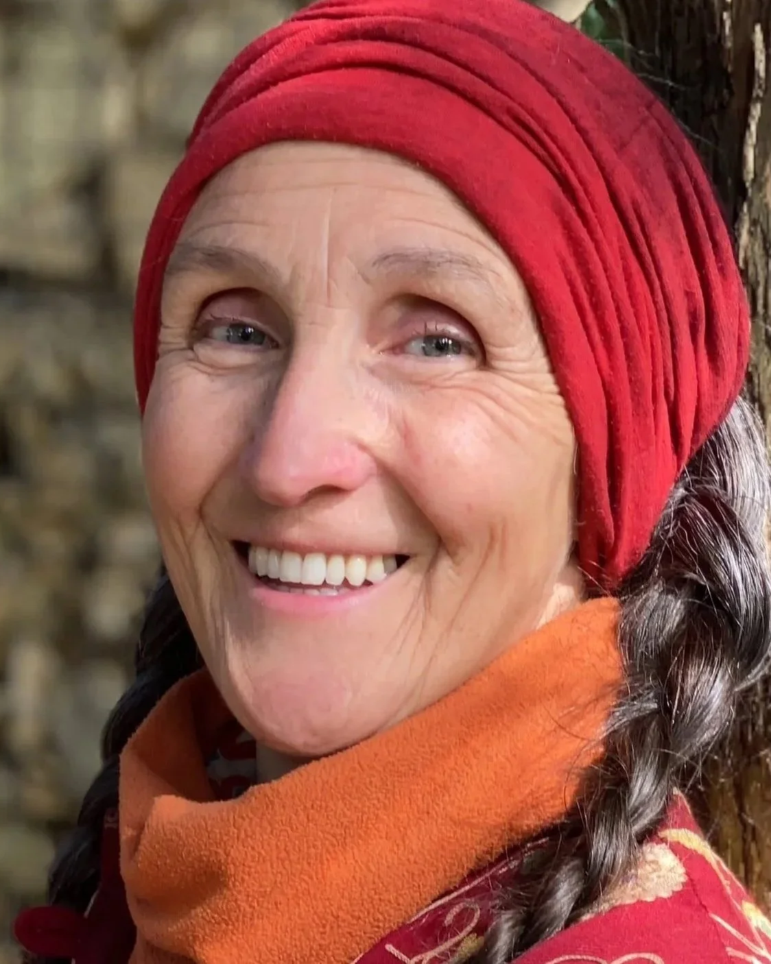 A smiling woman wearing a red headscarf, an orange scarf, and a red patterned jacket, standing outdoors next to a tree trunk.