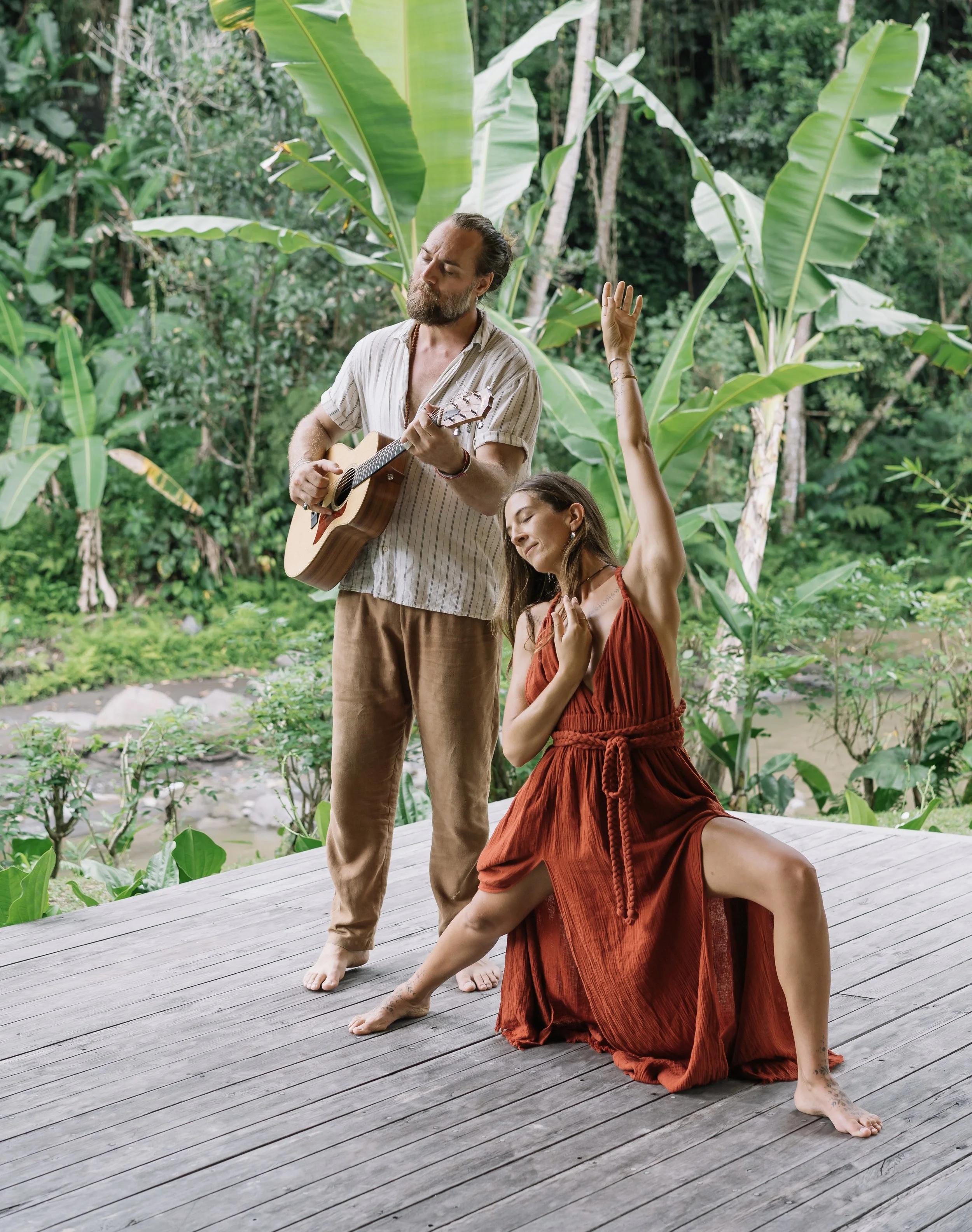 A man playing guitar and a woman in a red dress dancing outdoors on a wooden deck surrounded by lush green tropical plants.