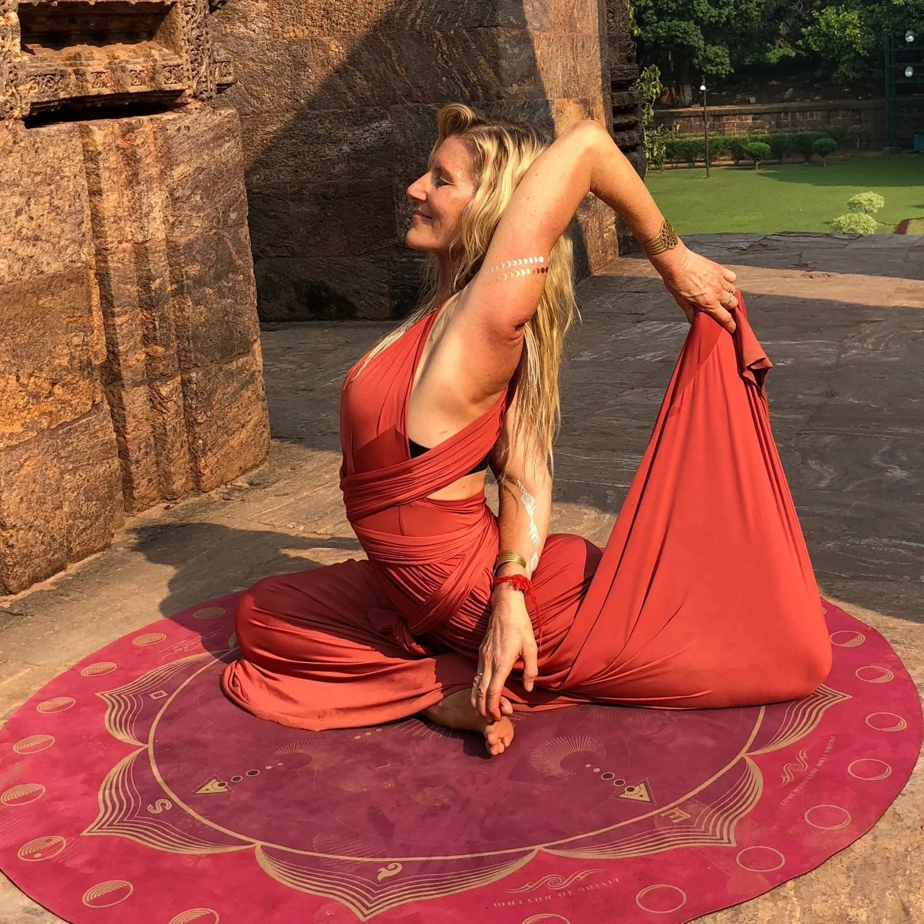 A woman practicing yoga outdoors on a red yoga mat, wearing a reddish-orange flowing dress, sitting cross-legged while holding her foot behind her head with one hand and stretching her arm upward. She has long blonde hair and a serene expression.