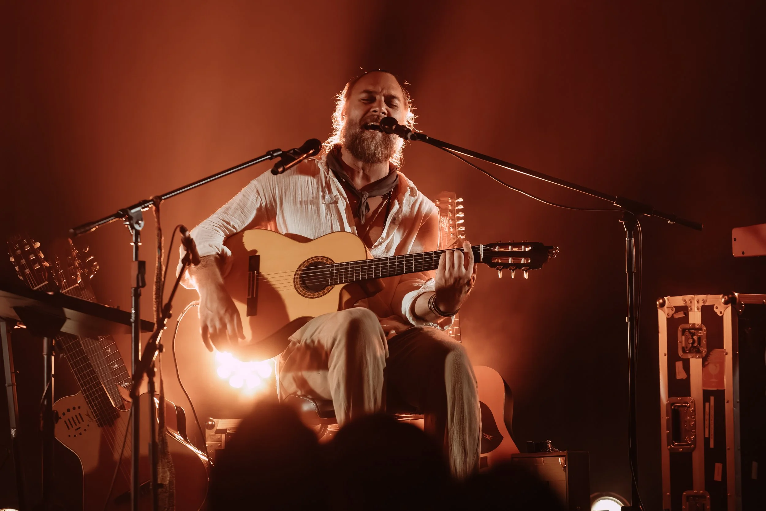 A man with a beard and long hair, singing passionately and playing an acoustic guitar on stage, surrounded by musical equipment and warm orange lighting.