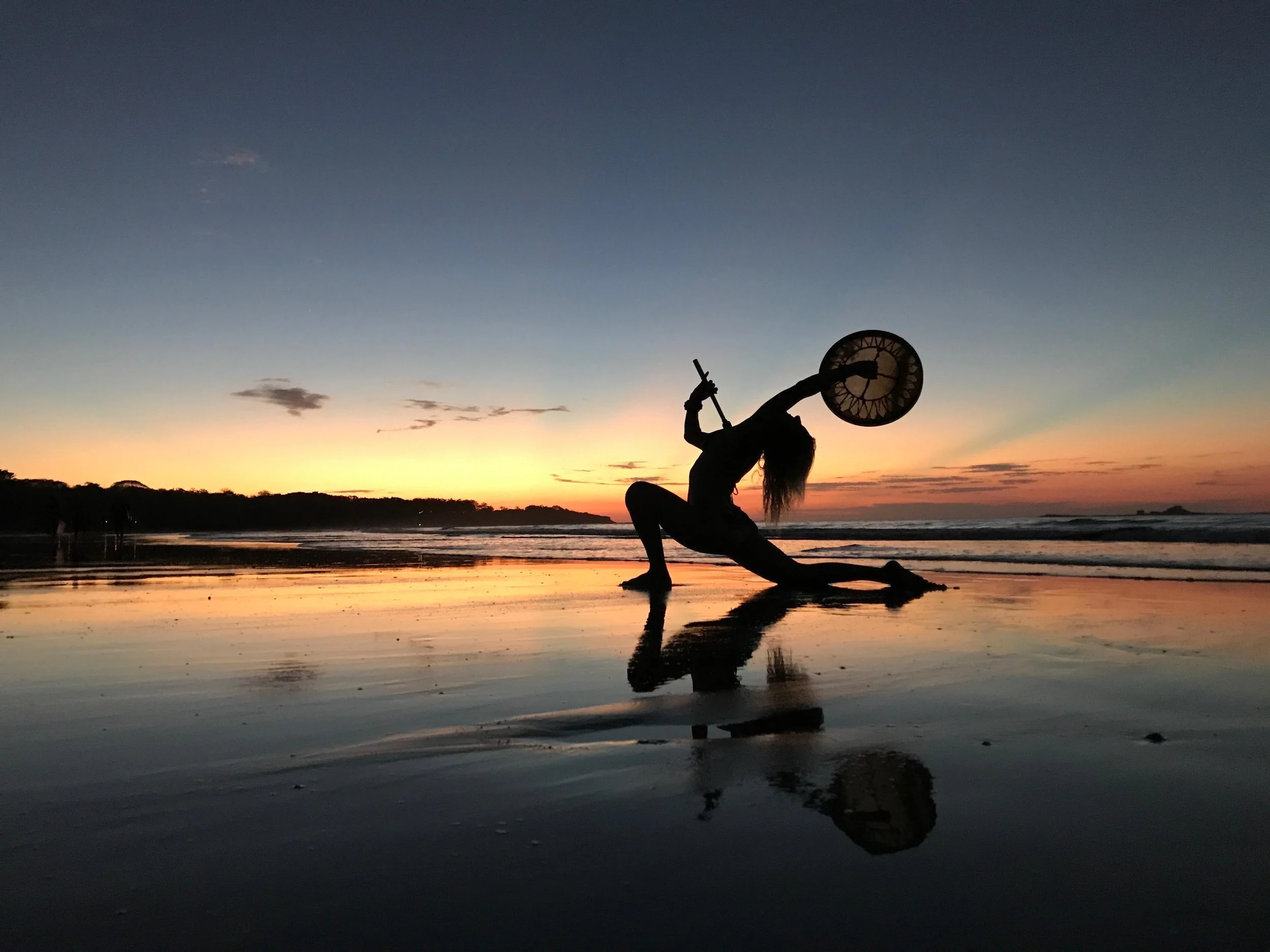 Silhouette of a woman practicing swordsmanship with a shield on a beach at sunset, with reflections in the wet sand and a colorful sky.