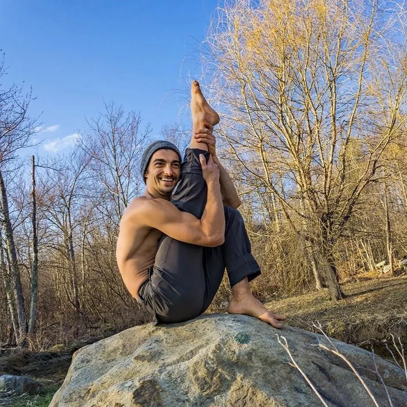 A shirtless man smiling, wearing a beanie, sitting on a large rock while holding his legs in a twisted yoga pose outdoors during fall with leafless trees in the background.