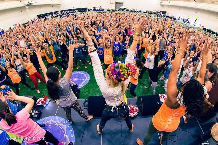 Large indoor event with many women participating in a group exercise or dance, led by an instructor in colorful attire, with a stage and microphones in the foreground.