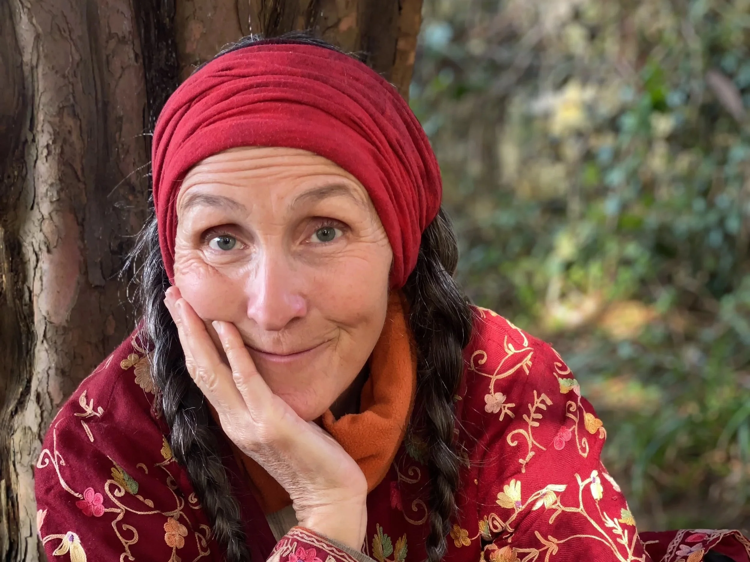 A woman with light skin, gray eyes, and long dark braided hair, wearing a red headscarf and a red embroidered top, resting her head on her hand and smiling while sitting outdoors with trees and greenery in the background.