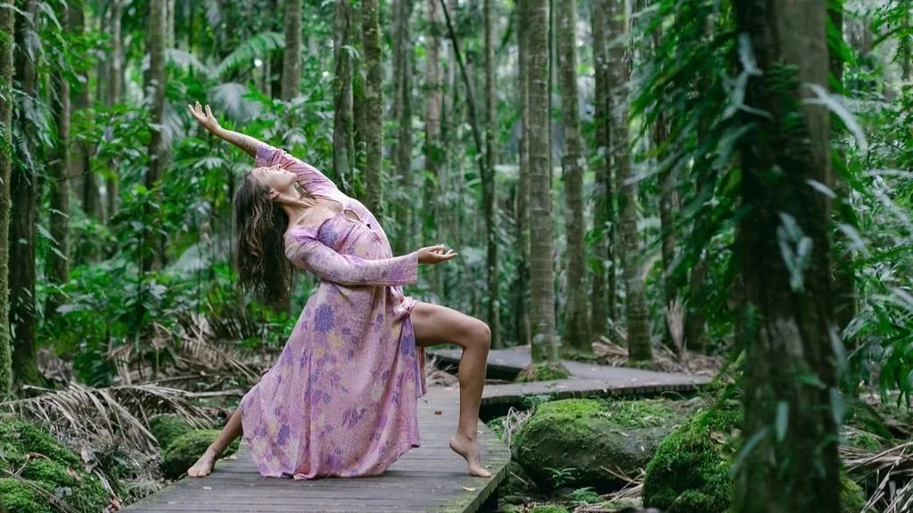 A woman in a flowing pink and purple dress striking a dance pose on a wooden pathway in a lush, green rainforest.